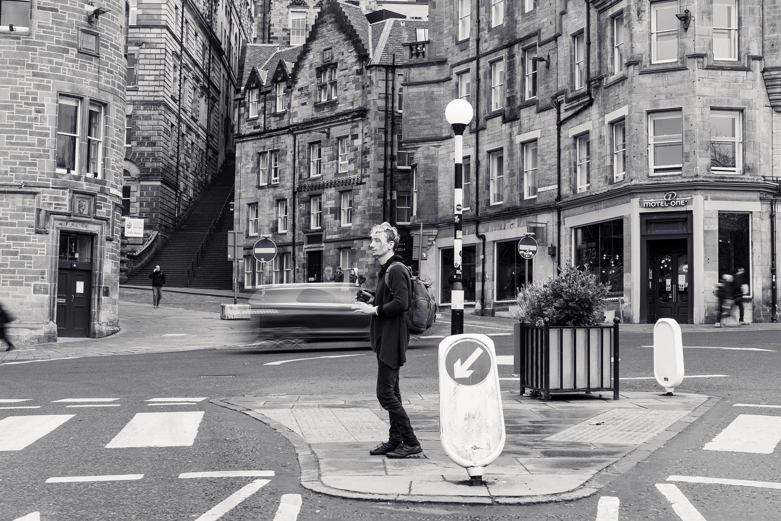 A young person with a backpack standing on a city crosswalk, holding a phone, with blurred cars passing by. The scene is in black and white, with old brick buildings in the background.