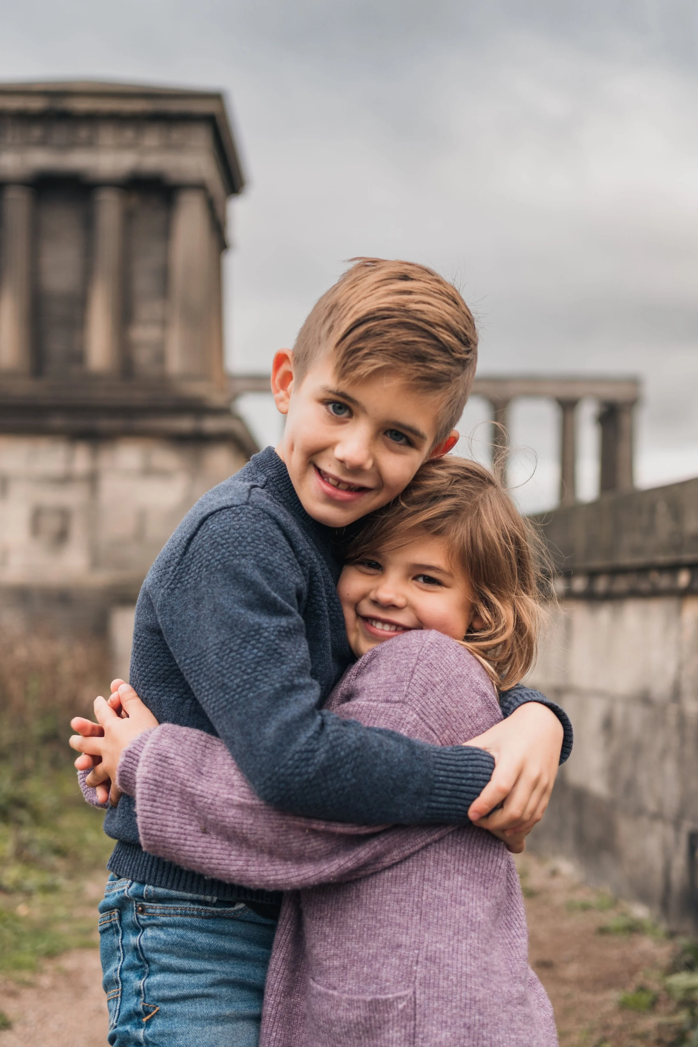 A boy and girl hugging outdoors on a cloudy day with a historical building in the background.