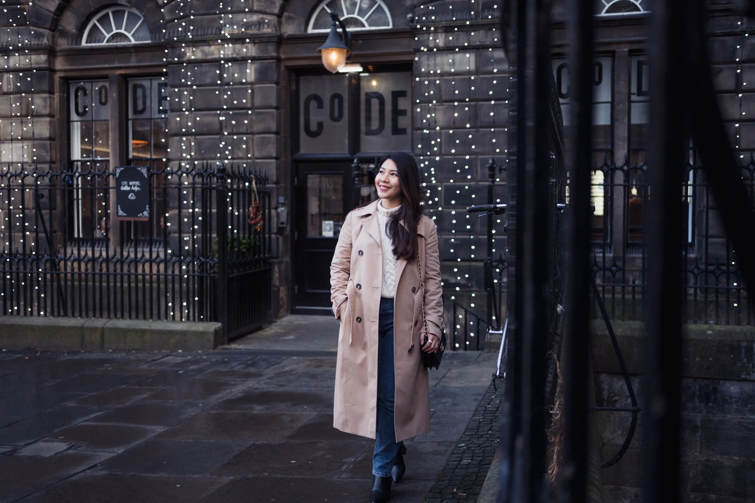 A woman walking on a city sidewalk during evening hours, smiling, wearing a beige trench coat, blue jeans, and black boots, with decorative string lights on a dark brick building in the background.
