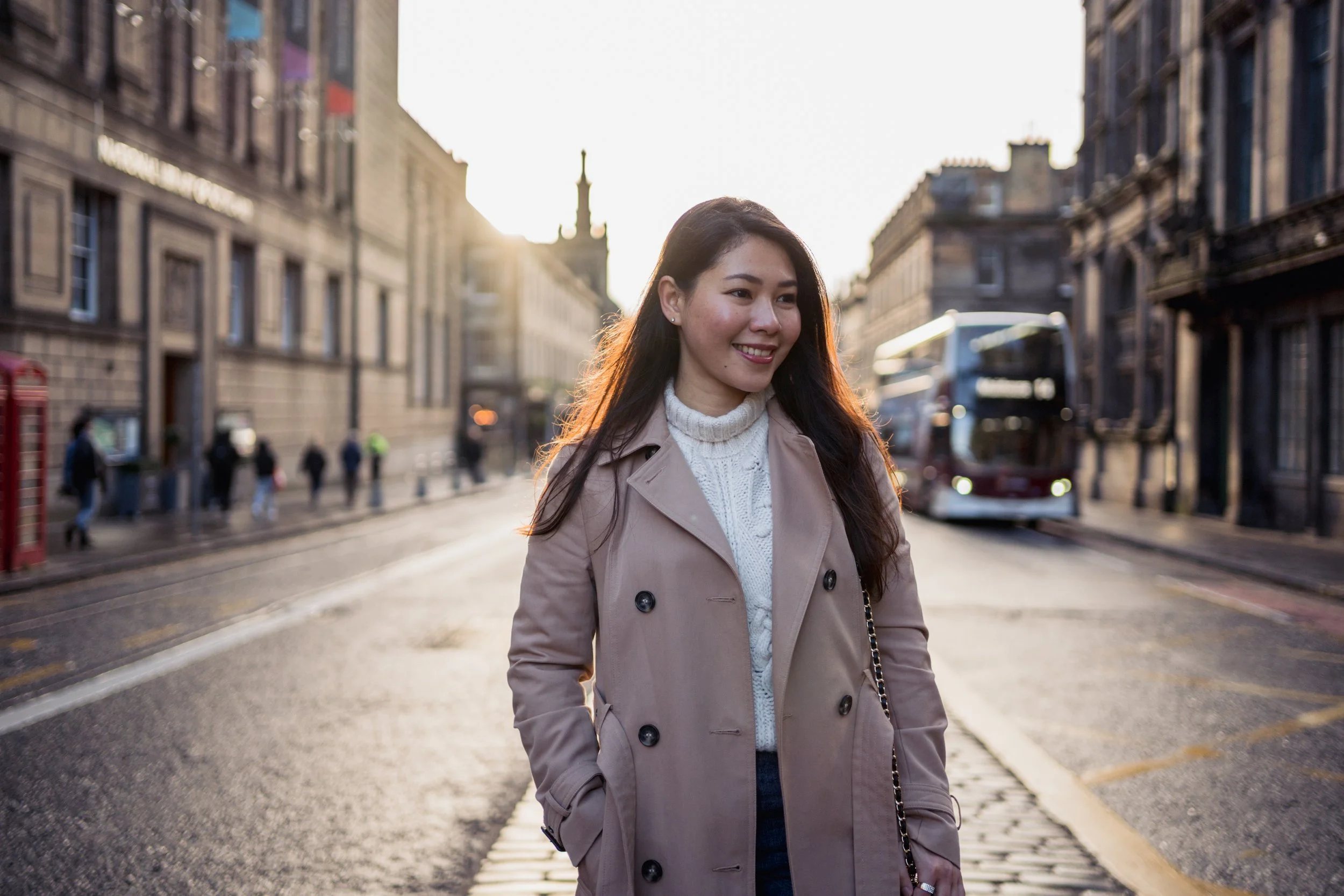 A woman with long dark hair smiling while standing on a city street at sunset. She is wearing a beige trench coat over a white knit sweater. In the background, there are buildings, pedestrians, and a red double-decker bus.