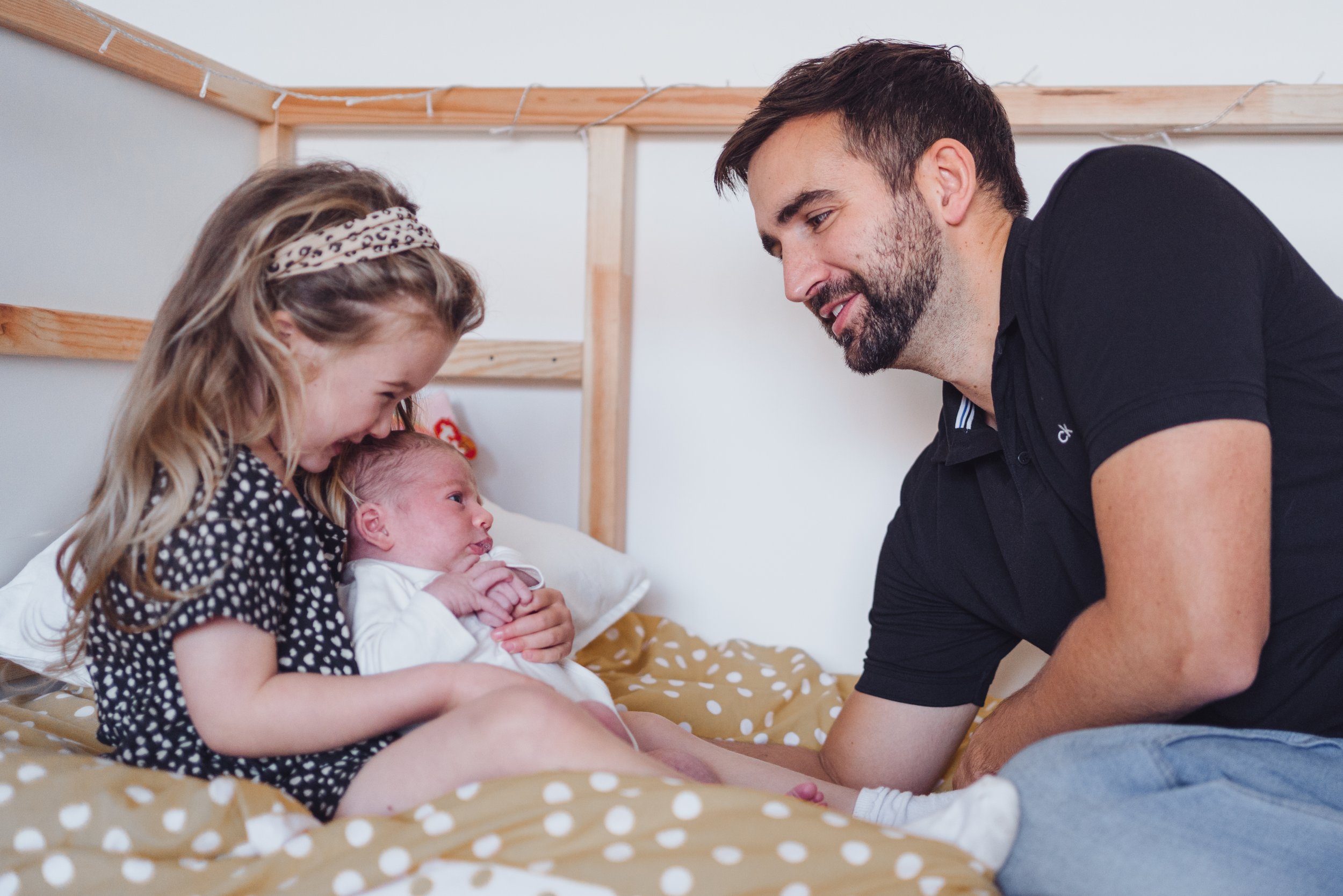 A young girl, a newborn baby, and a man in a bedroom. The girl and the man are holding the baby and smiling.