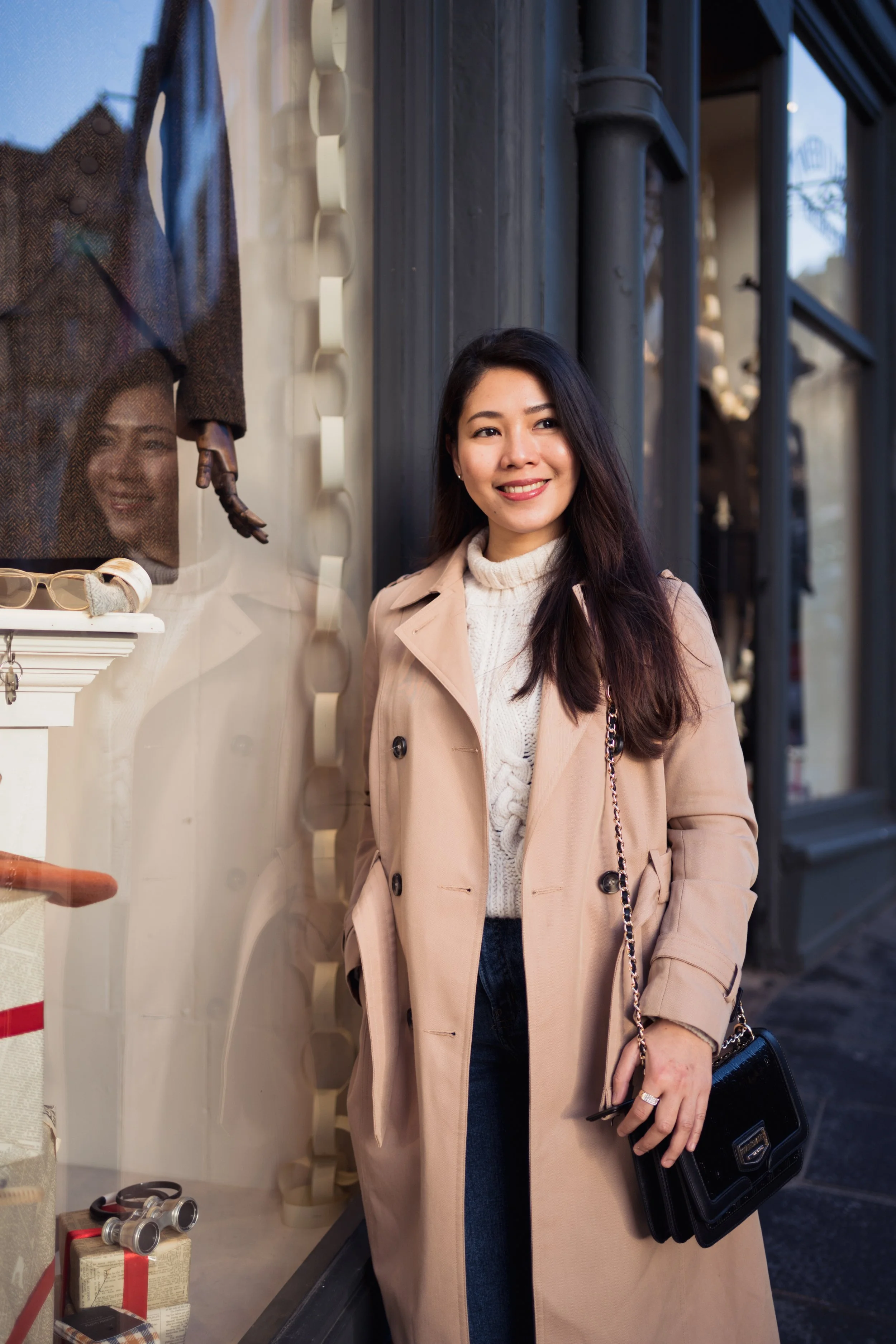 A woman stands outside a storefront window, smiling, wearing a beige trench coat, white turtleneck sweater, and holding a black purse.