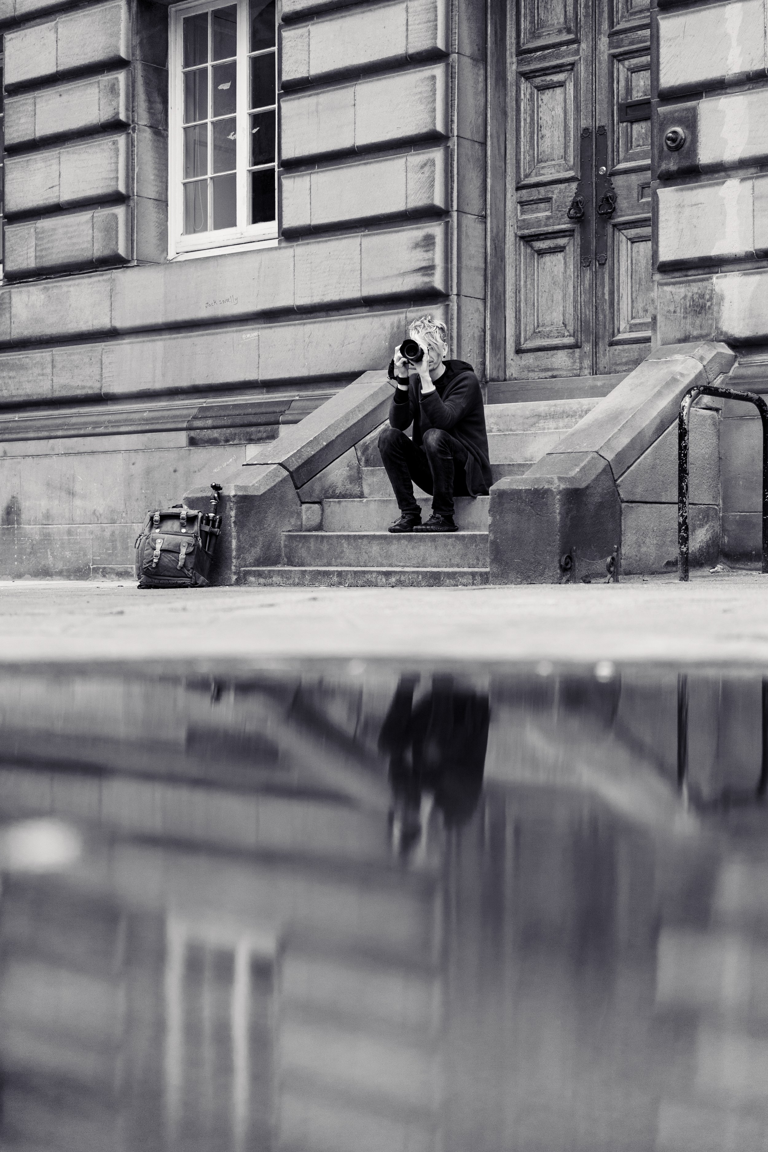 A person sitting on stone steps outside a building, taking a photograph with a camera. A backpack is on the ground nearby, and the building features a large wooden door and a window. The photo is black and white.