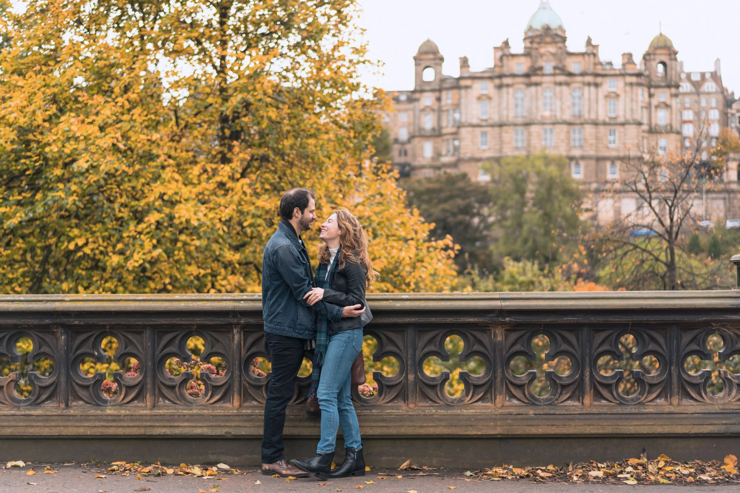 A couple standing close together near a decorative bridge railing in autumn, smiling at each other with a cityscape and colorful fall trees in the background.