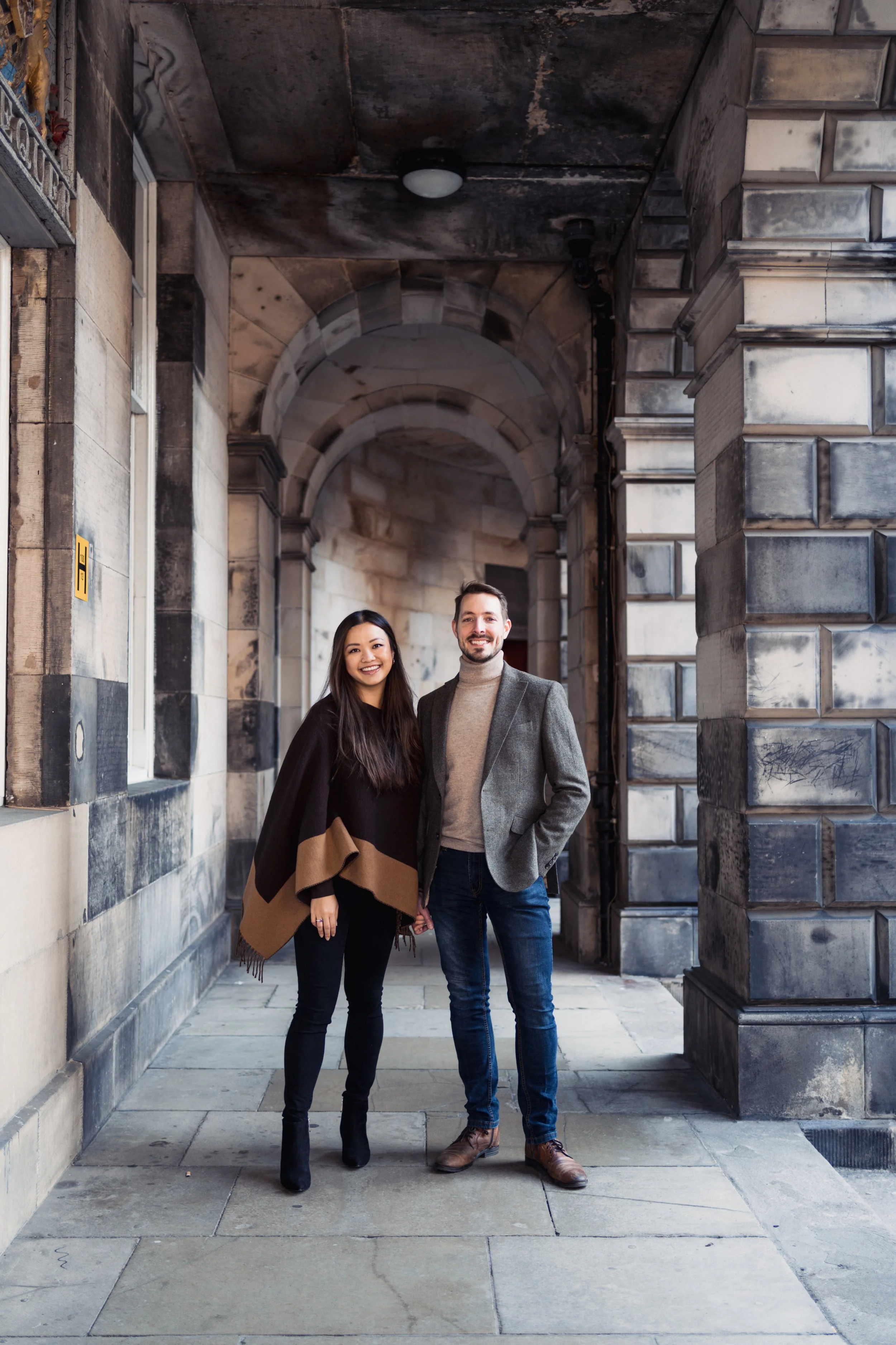 A couple smiling and holding hands, standing in front of a historic stone building with archways and detailed architecture.