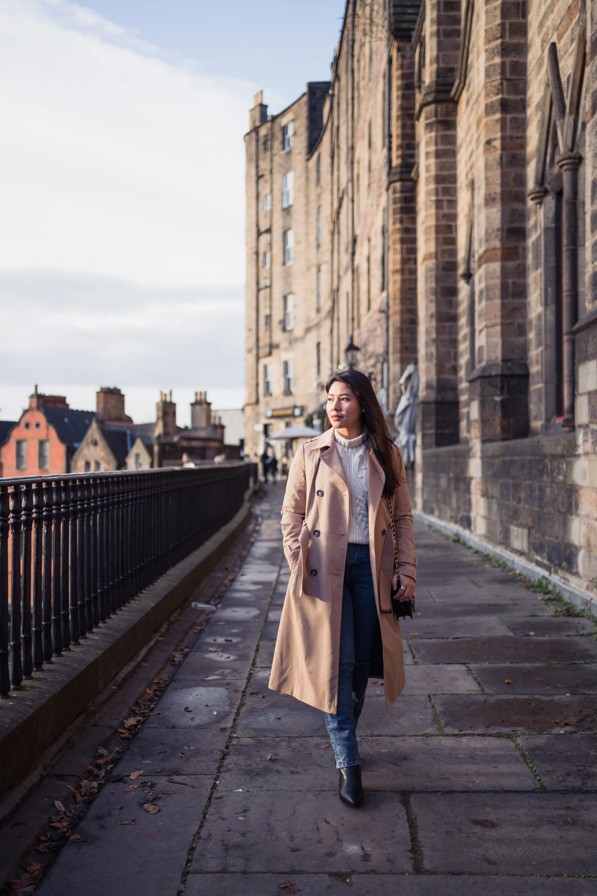 A woman walking on a stone-paved sidewalk beside historic brick buildings, wearing a beige trench coat, jeans, and black boots, with the sky partly cloudy.