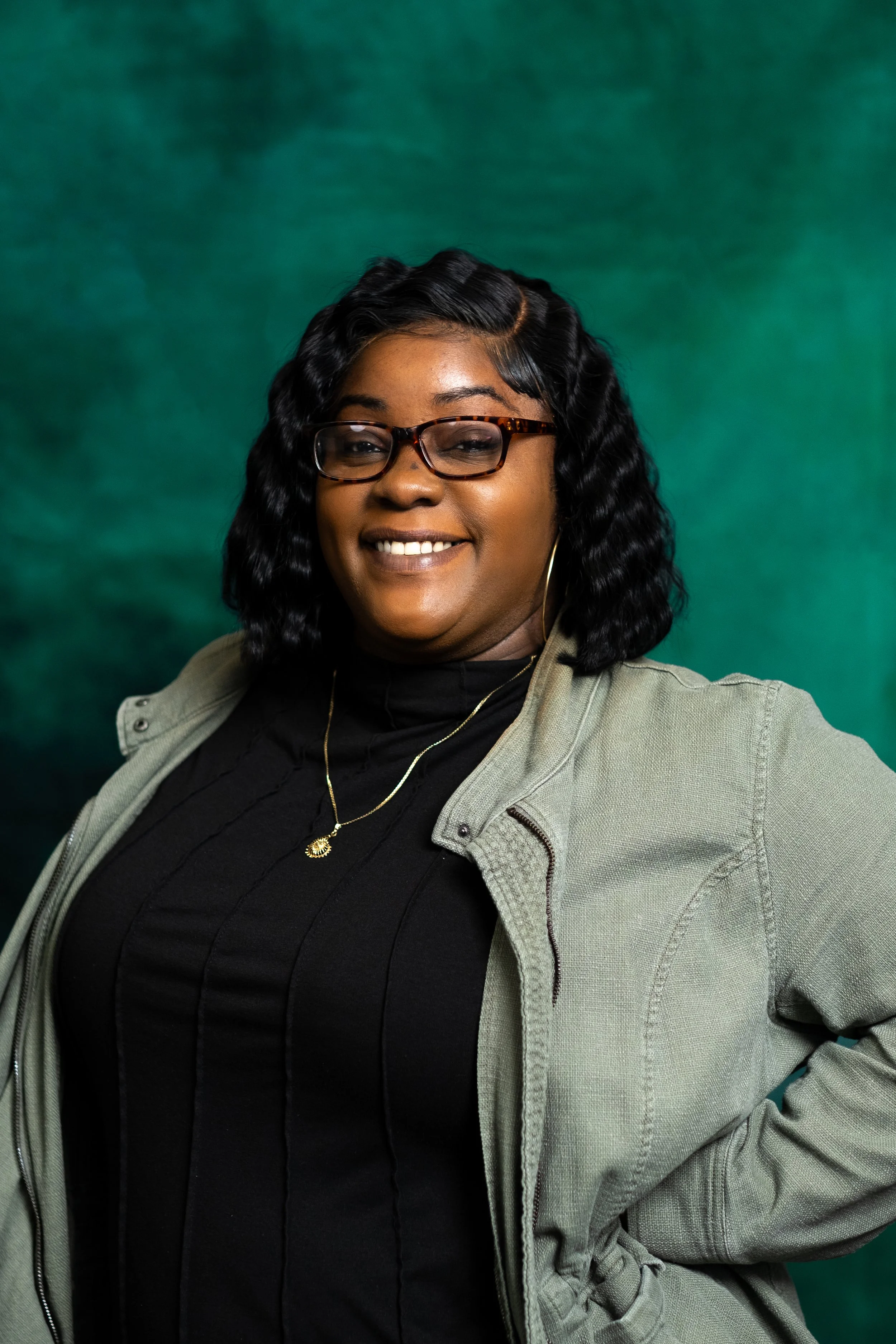 A woman with wavy dark hair, glasses, and hoop earrings smiling at the camera, wearing a graphic t-shirt and a black jacket against a green background.