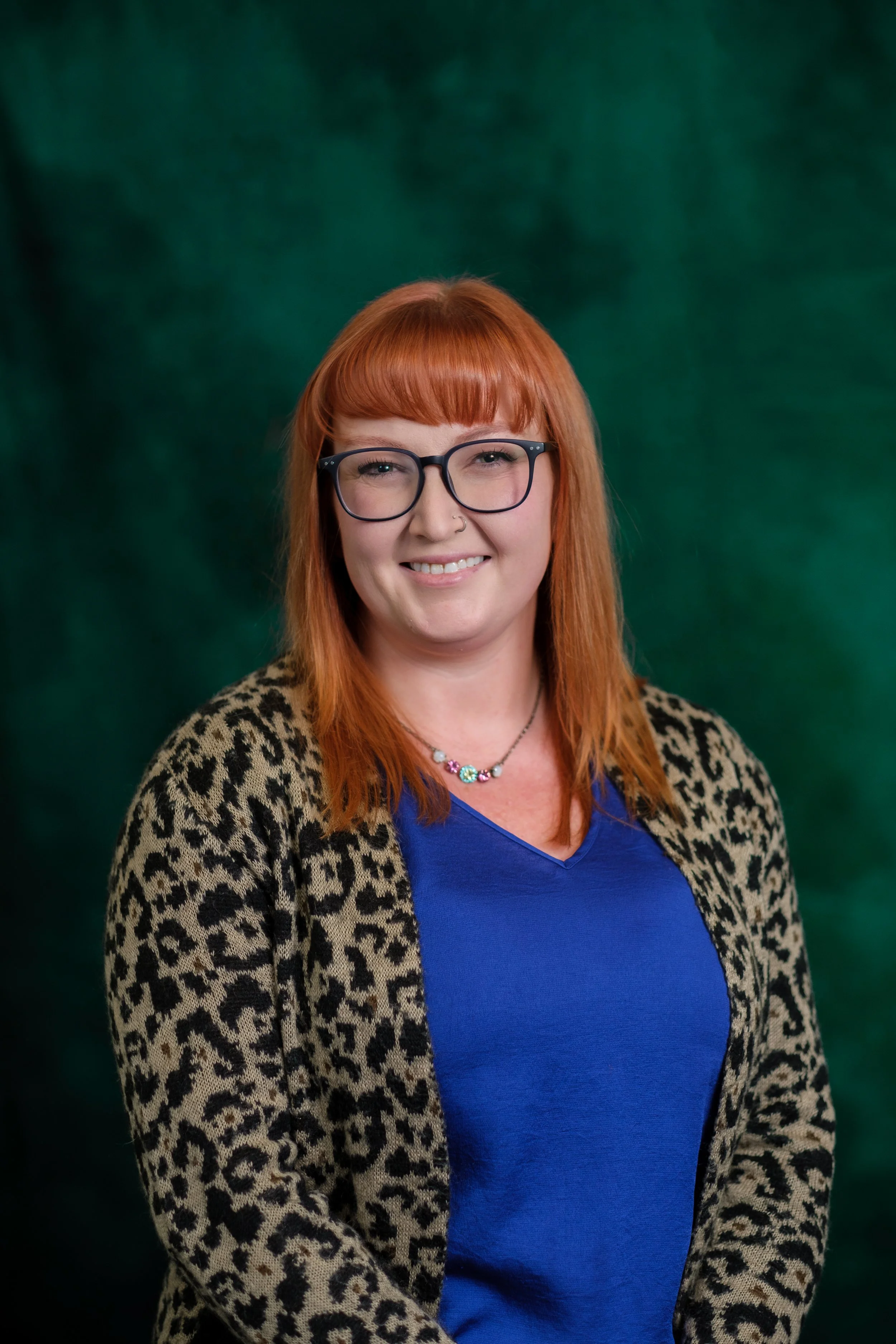 A woman with red hair, wearing a blue V-neck top, layered pearl and chain necklaces, and smiling against a green background.