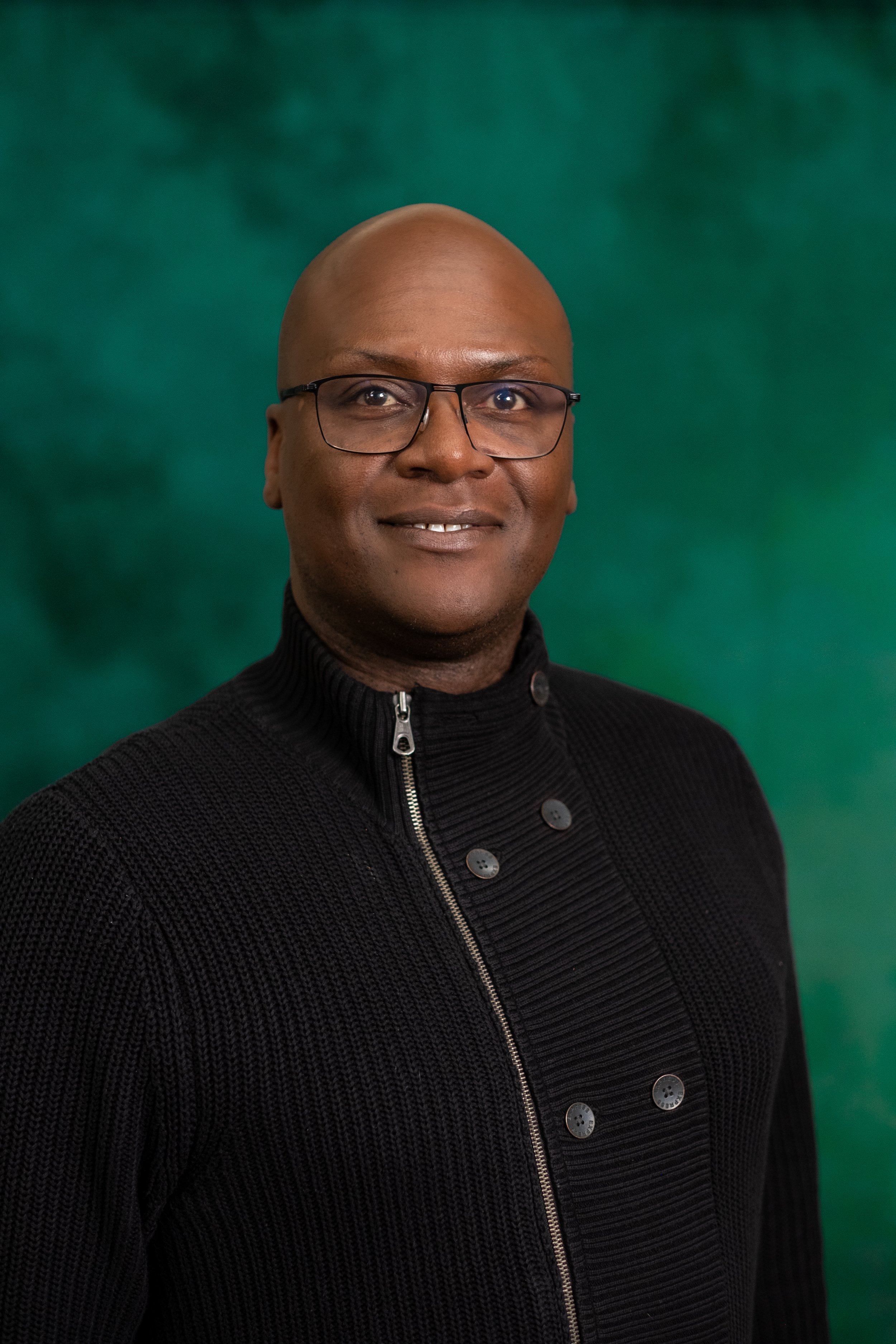 A smiling man wearing glasses and a black and white checkered shirt against a green background.