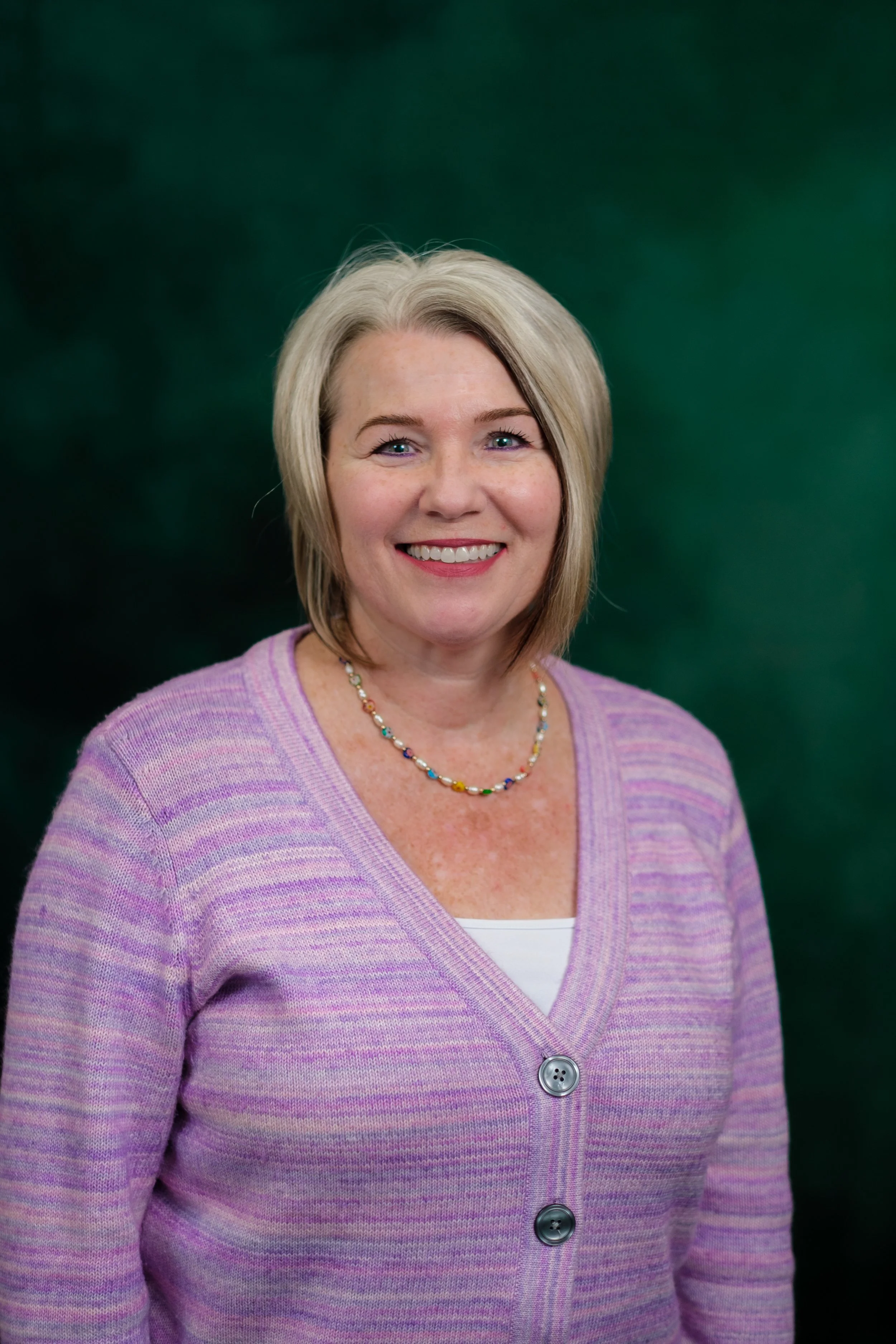 A smiling woman with blonde and gray hair, wearing a brown patterned cardigan and a pearl necklace, standing against a green background.
