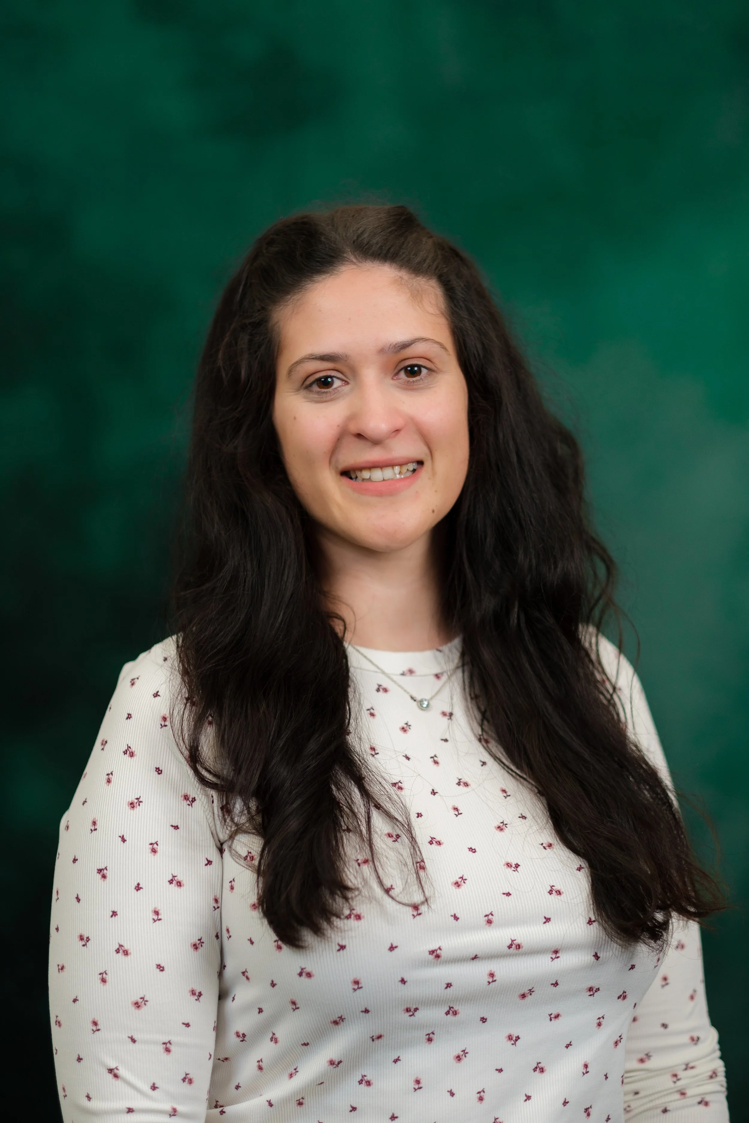 A young woman with long, dark wavy hair, smiling, wearing a mustard yellow sweater, standing in front of a green textured backdrop.