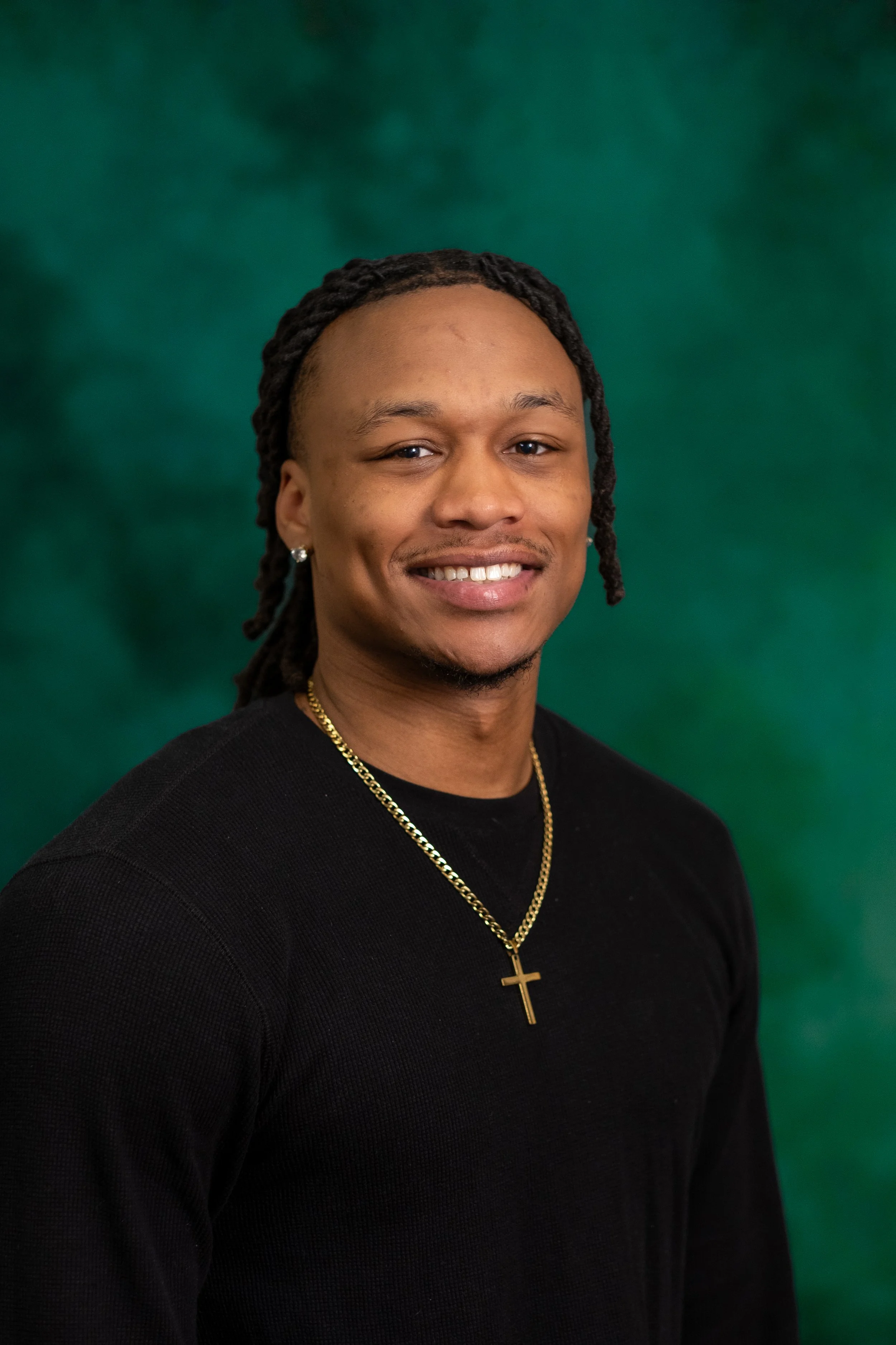 Portrait of a smiling young man wearing a plaid jacket and earrings, standing against a green background.
