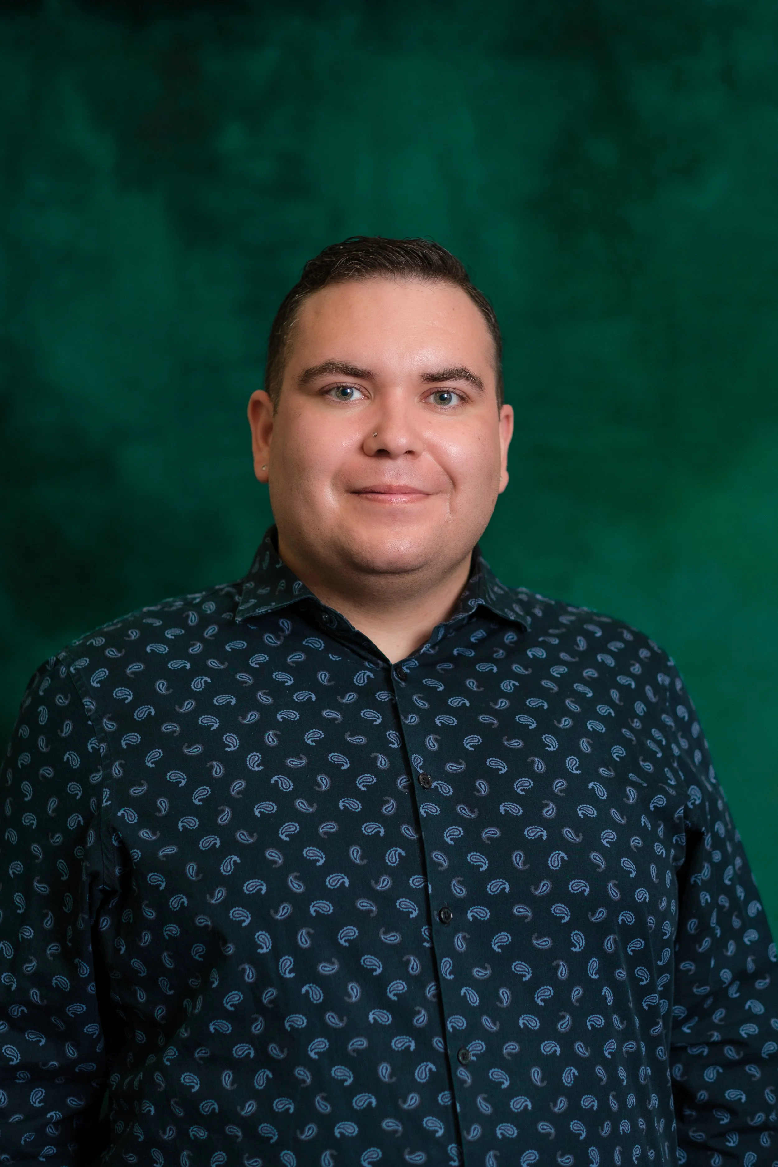 Portrait of a young man with short dark hair, wearing earrings, a black patterned shirt, and a gray T-shirt underneath, standing against a green backdrop.