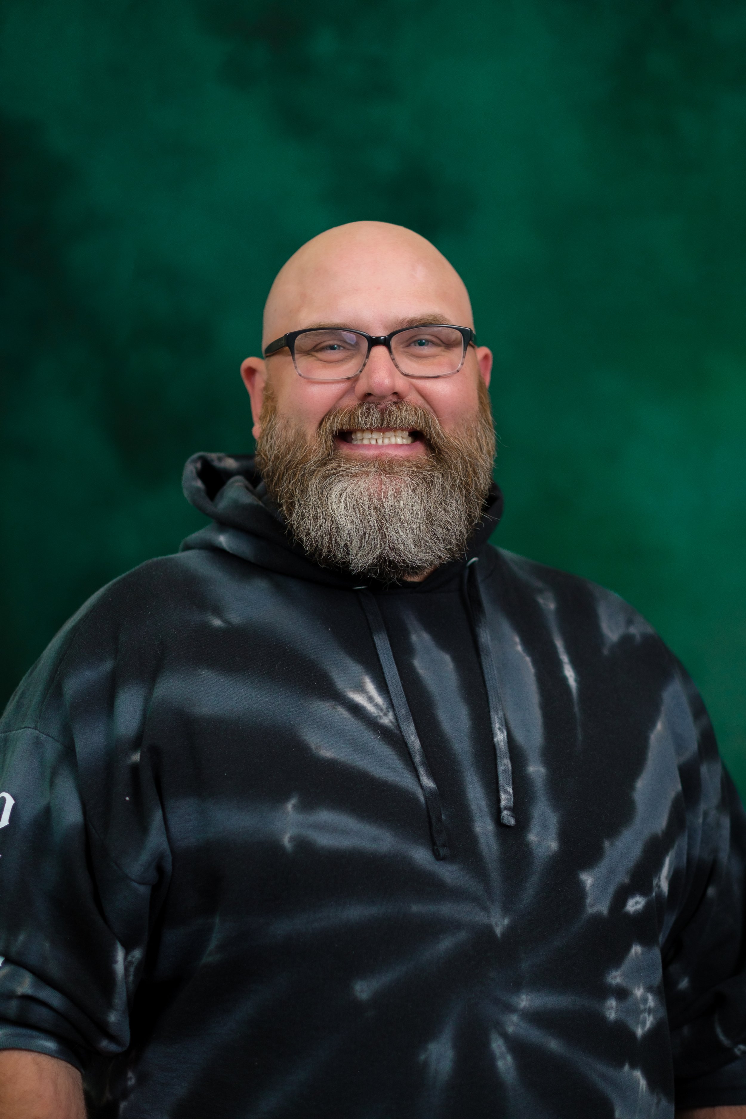 A smiling man with glasses, bald head, and a beard, wearing a black shirt, against a green blurred background.