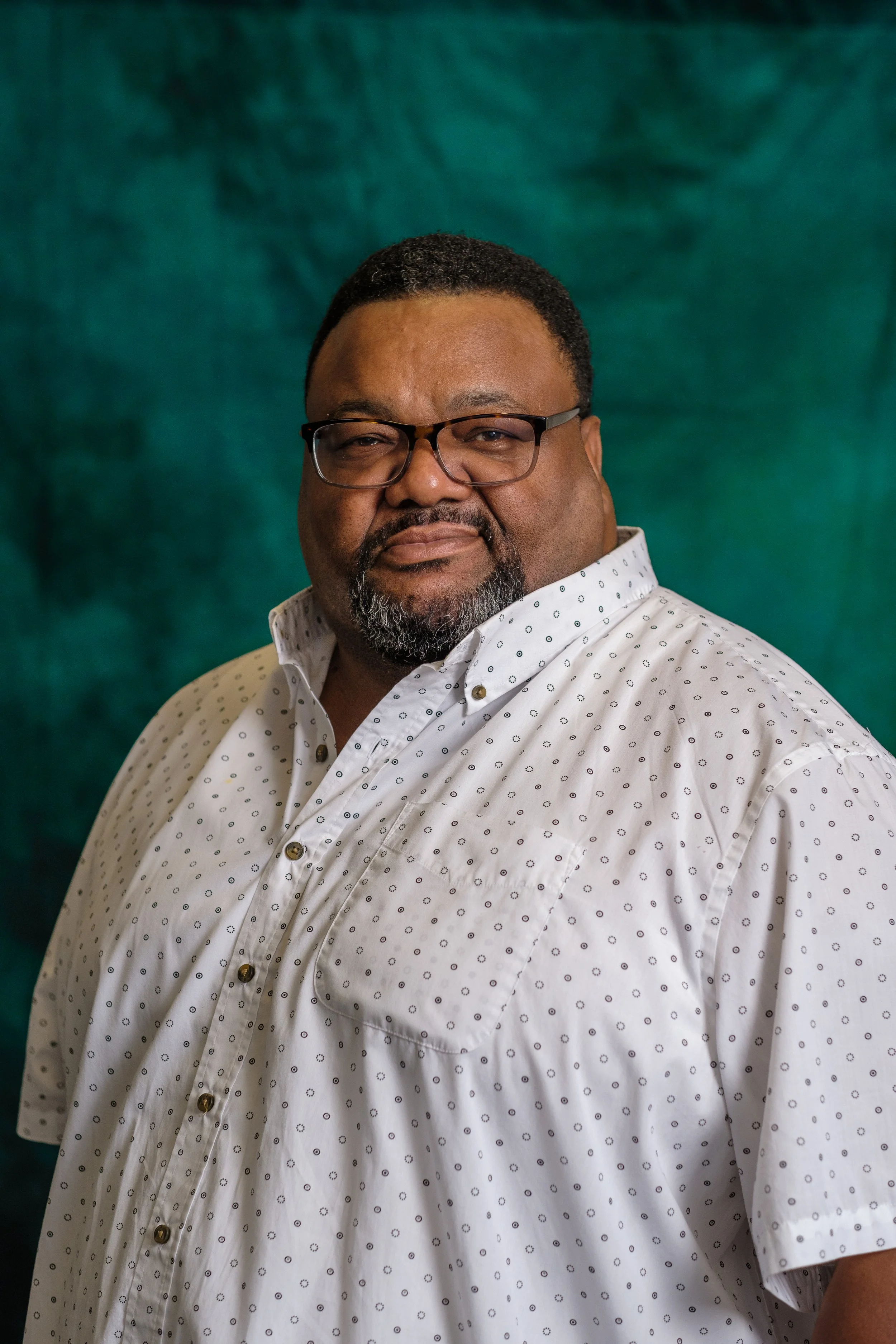 A man with glasses and a beard wearing a light green shirt stands against a green background.