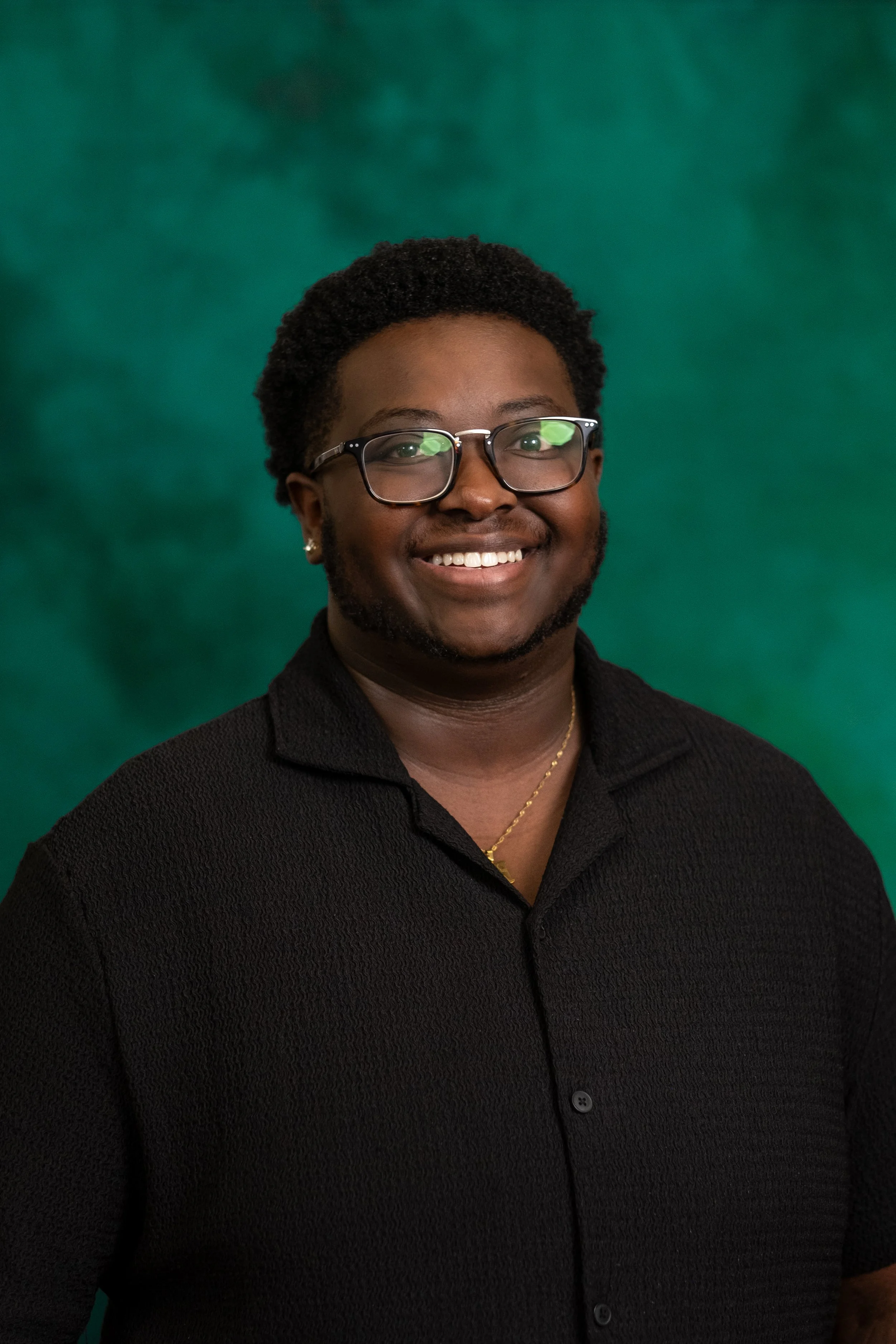 Portrait of a young person with short curly hair, wearing glasses, a purple corduroy jacket over a light-colored shirt, standing against a green background with a warm smile.