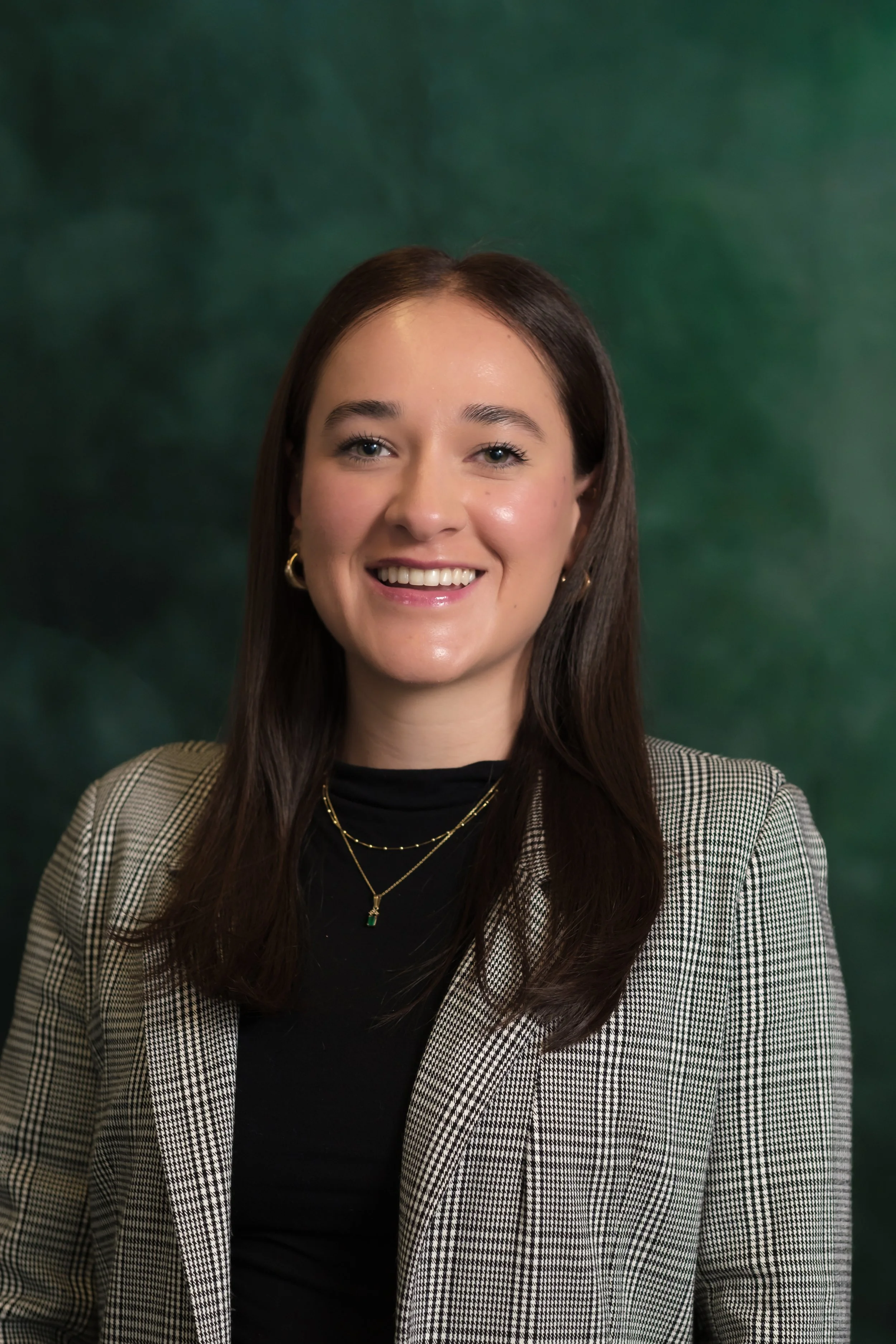 A woman with long brown hair, wearing a black turtleneck and gold jewelry, smiling in front of a green background.