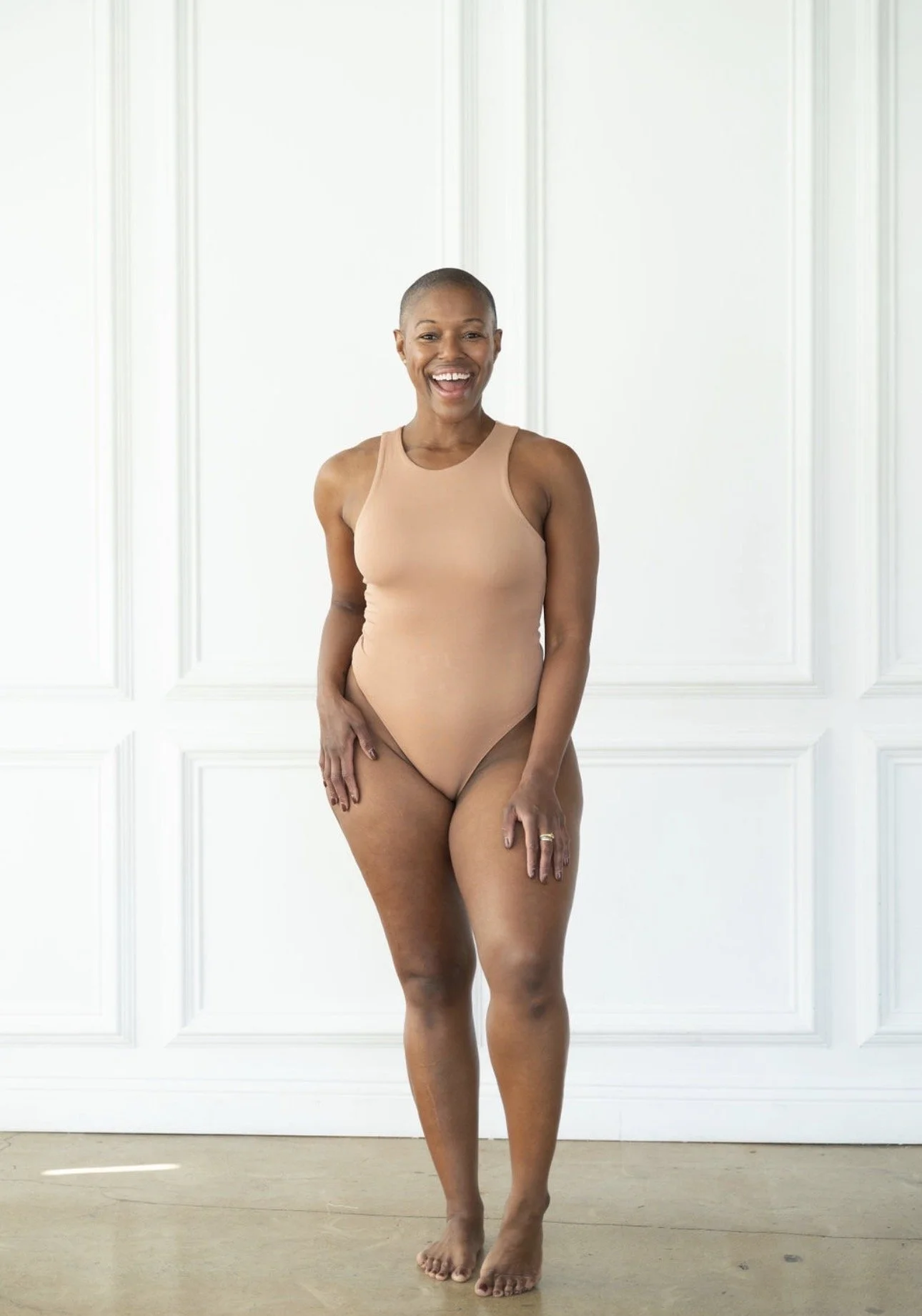 A woman with a shaved head wearing nude-colored underwear, standing in front of a white paneled wall, smiling and looking at the camera.