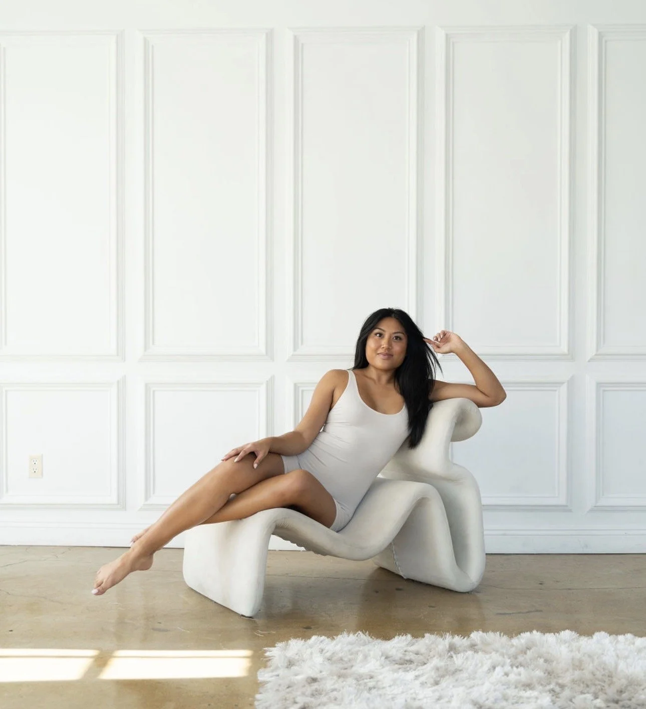 A woman in a white tank top and shorts reclining on an abstract white sculpture in front of a white paneled wall.