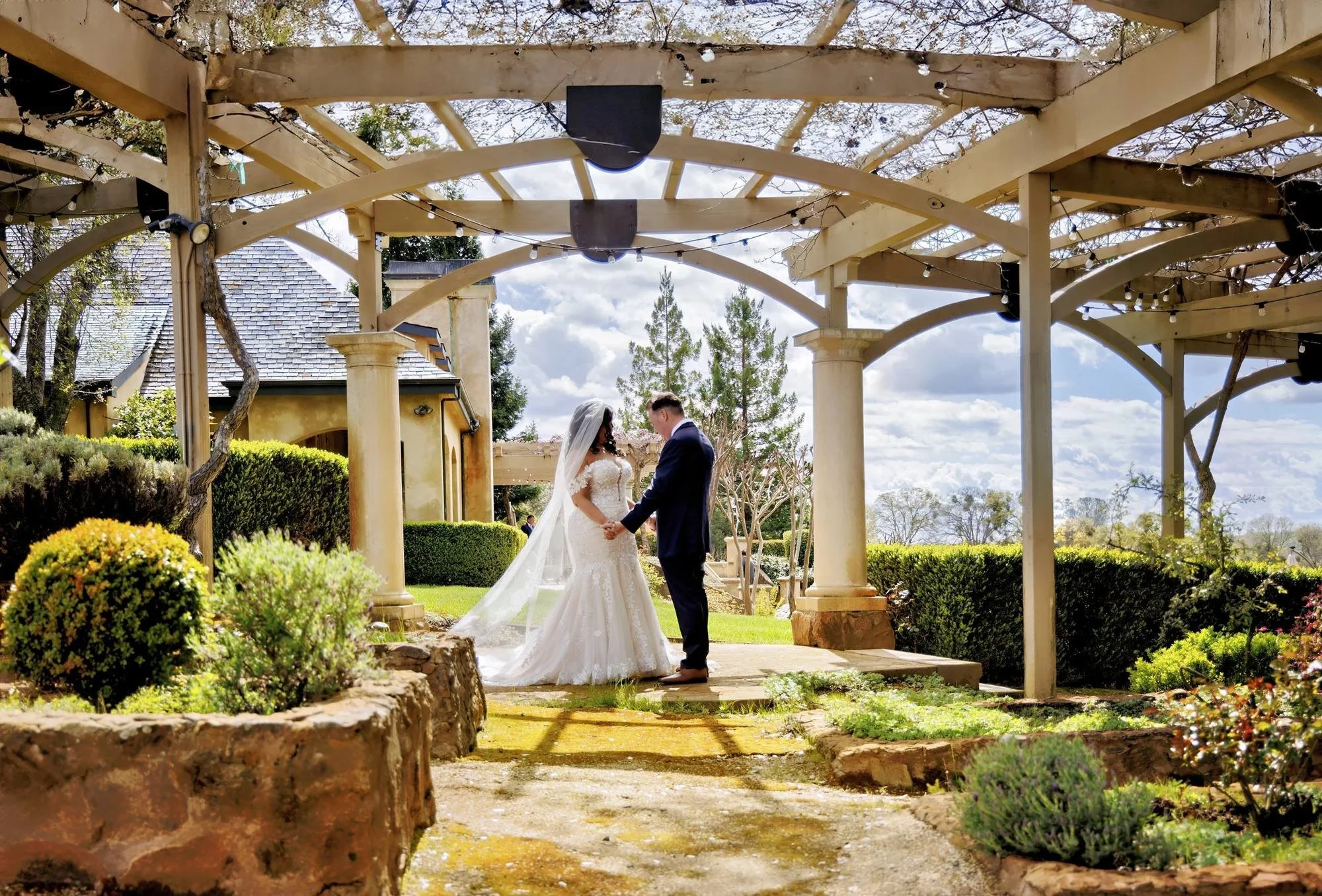 A bride and groom holding hands in an outdoor wedding ceremony under a wooden arbor, surrounded by landscaped greenery, with a partly cloudy sky above.