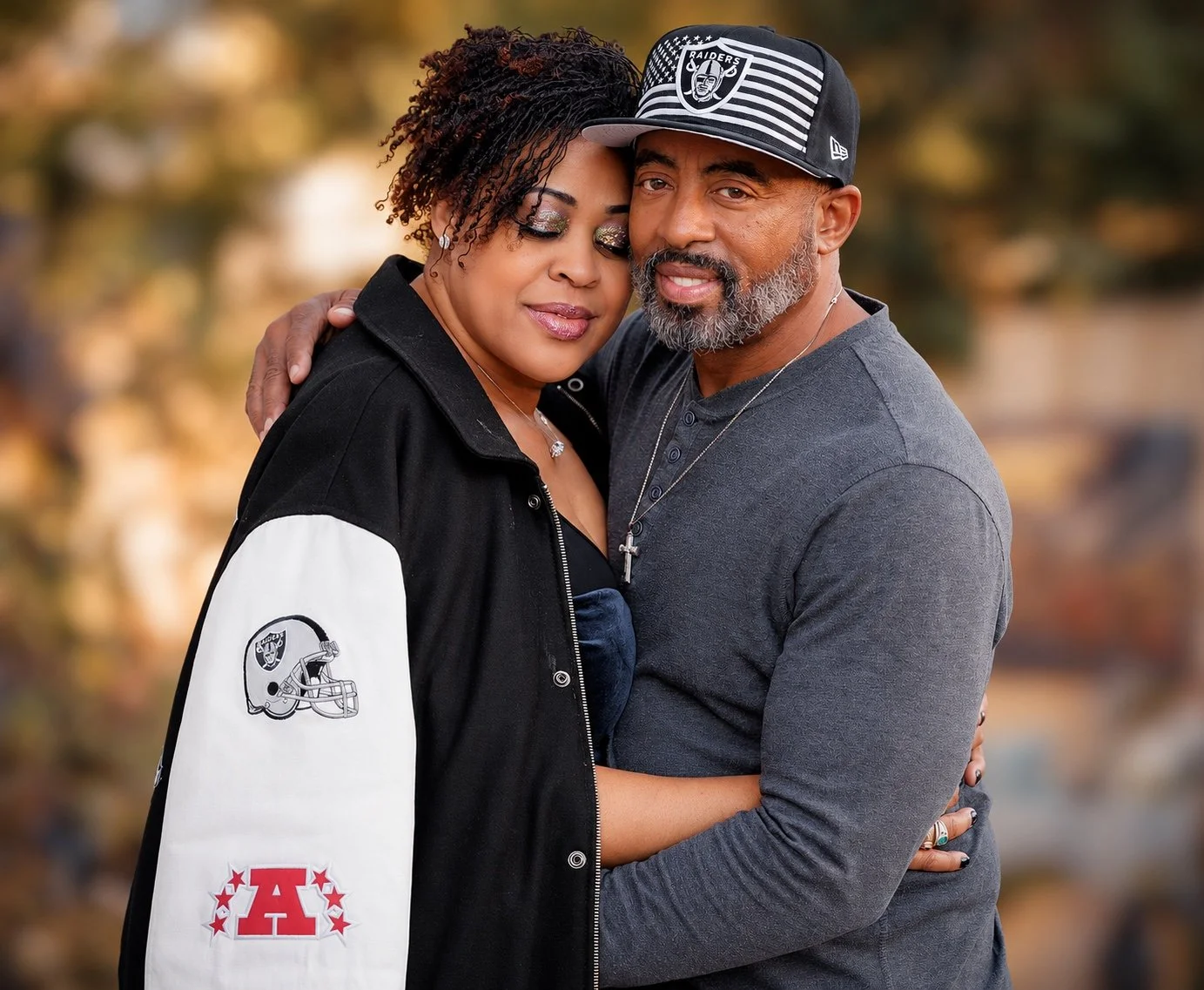 Couple embracing outdoors in Raiders apparel against a softly blurred golden background.