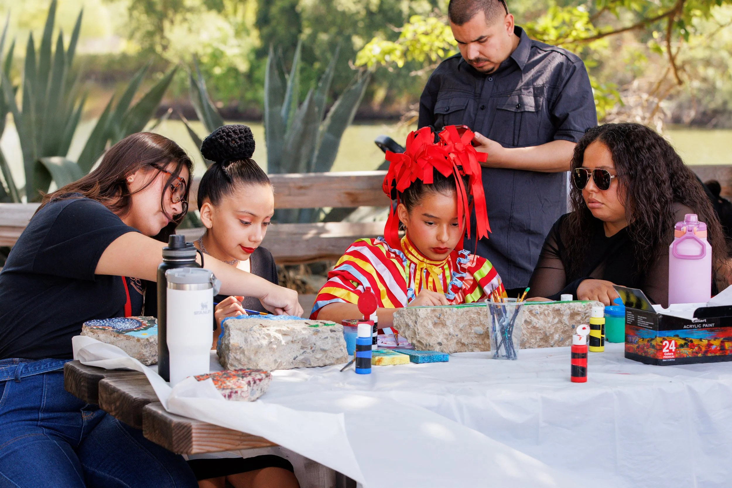Children and adults painting rocks at an outdoor table, with greenery in the background.