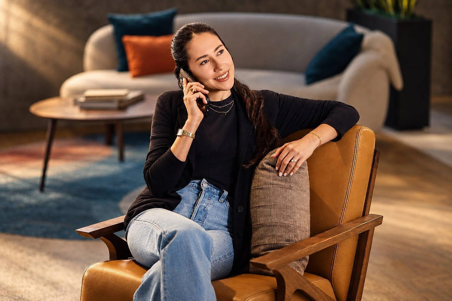 Woman sitting in a chair, talking on a mobile phone, smiling, in a cozy living room with a sofa and coffee table in the background.