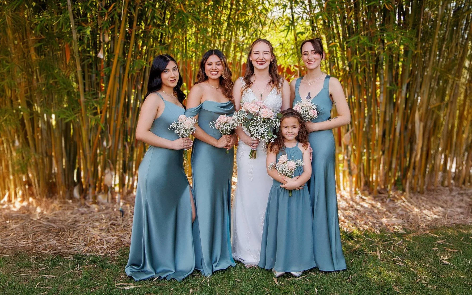Group of five women and one young girl in formal dresses standing outdoors in front of tall green plants, holding bouquets of flowers, during a wedding or special event.