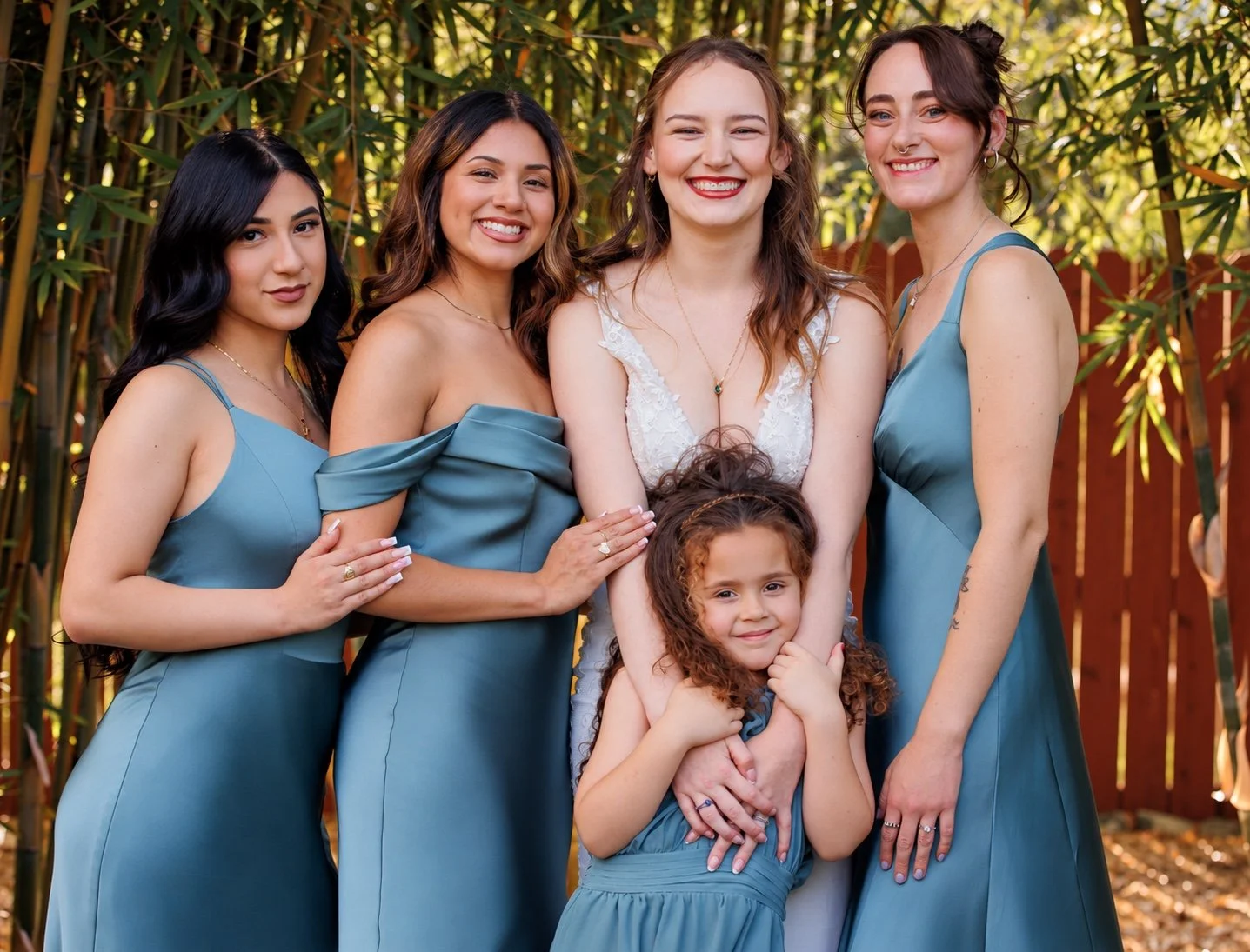 Bride with three bridesmaids and a young flower girl in teal dresses, posing together in front of bamboo.