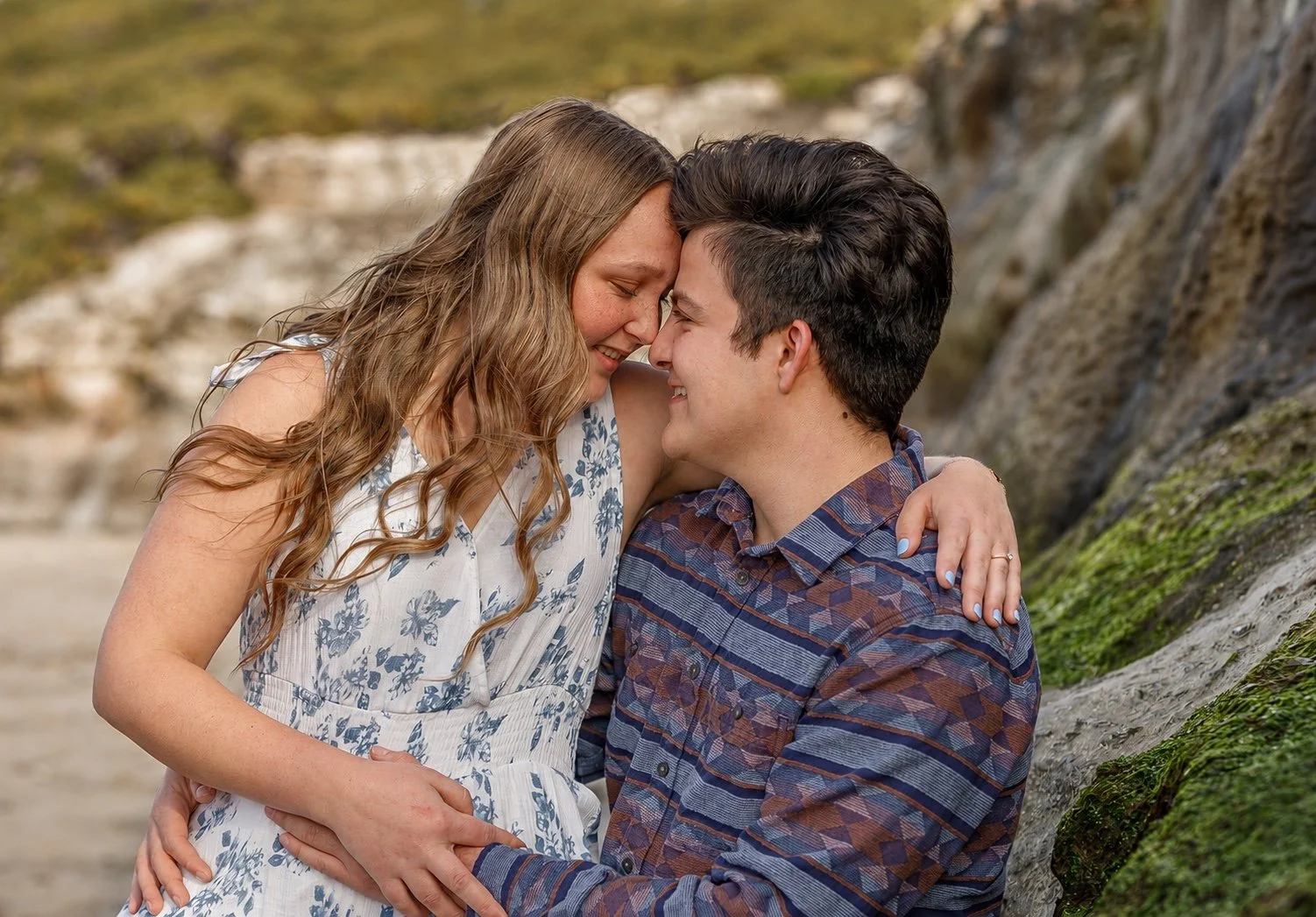 Couple embracing beside a moss-covered rock, smiling with their foreheads touching.
