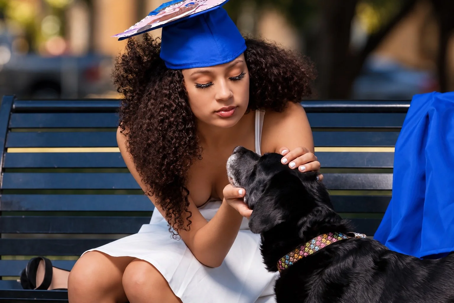 Graduate in a blue decorated cap and white dress petting a black dog while seated on a park bench.