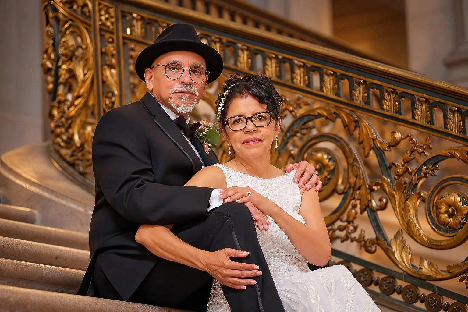 Bride and groom seated together beside an ornate gold staircase railing.
