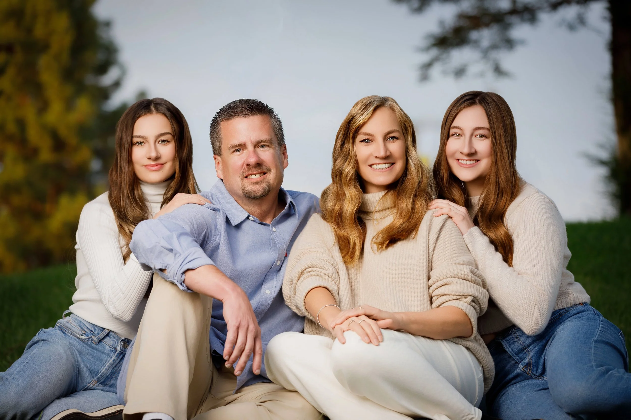 A family of four sitting outdoors on grass, with trees in the background. The father is in the center, with two daughters on either side, and a mother sitting in front of the father. They are smiling and dressed casually.