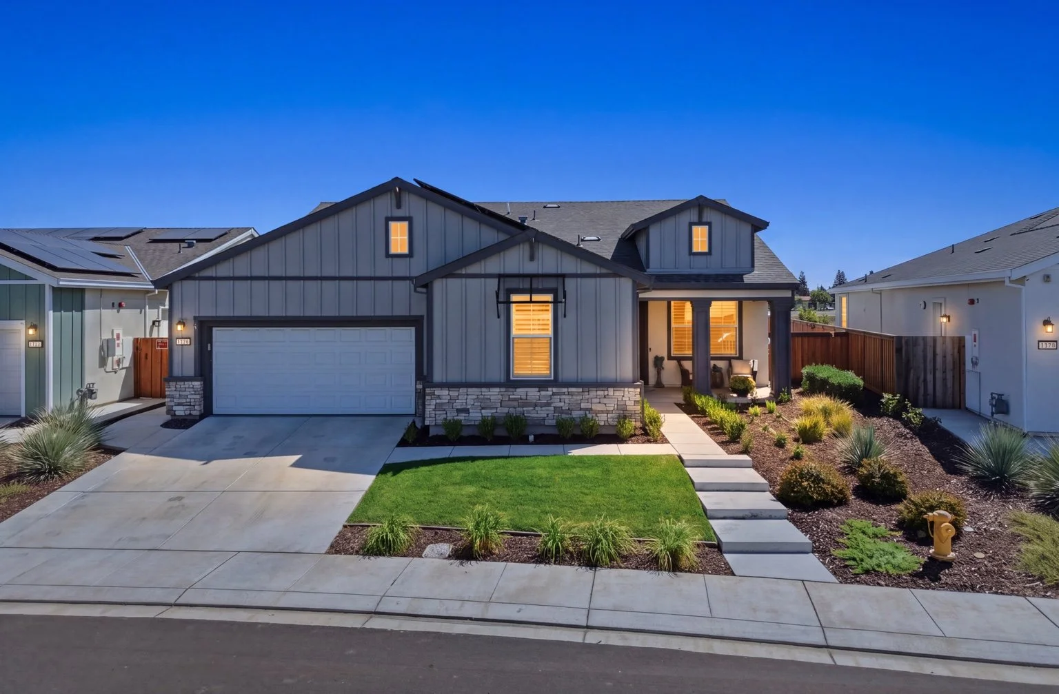 Gray modern farmhouse at twilight with warm window light, a landscaped front yard, and a blue evening sky.