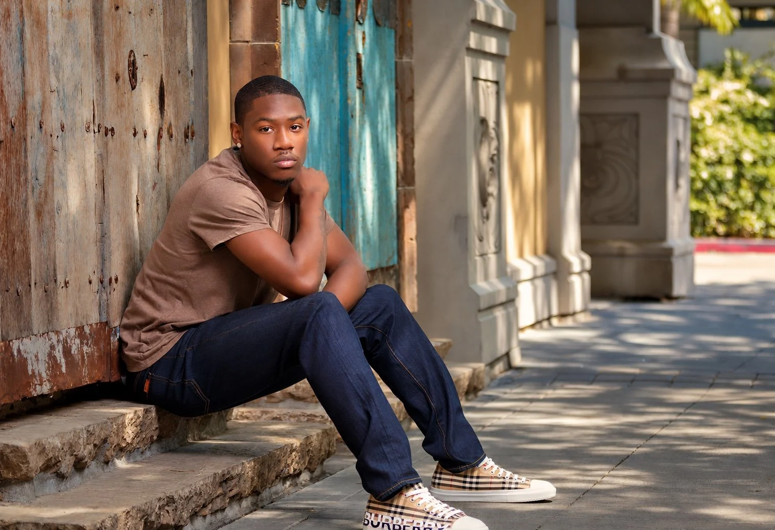 Man in a brown shirt and jeans seated on stone steps beside weathered wooden and turquoise doors.