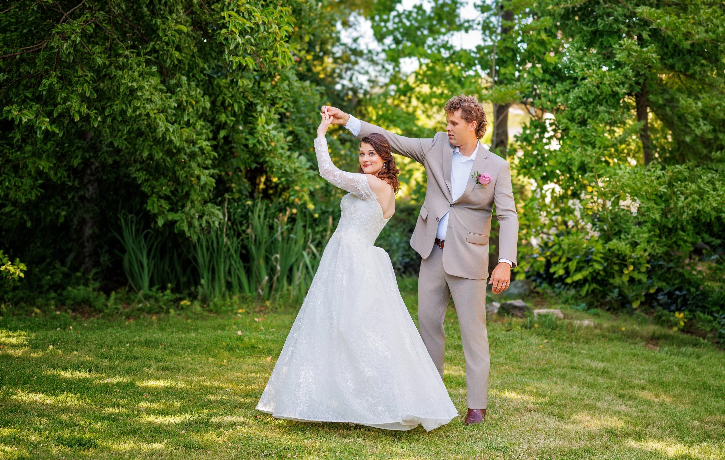 A bride and groom are dancing outdoors in a garden, surrounded by green trees and grass. The bride wears a white lace wedding dress, and the groom wears a light gray suit with a pink boutonniere. They are holding hands and looking at each other.