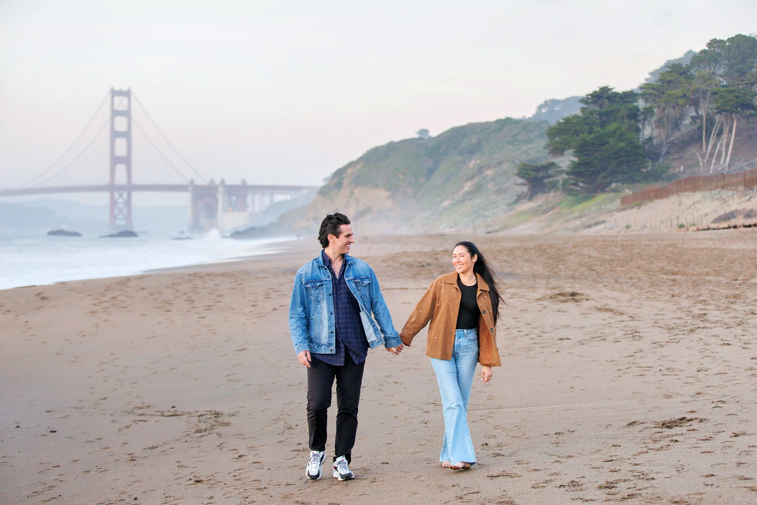 A young man and woman holding hands and walking on a beach with the Golden Gate Bridge in the background, smiling and enjoying the scenery.