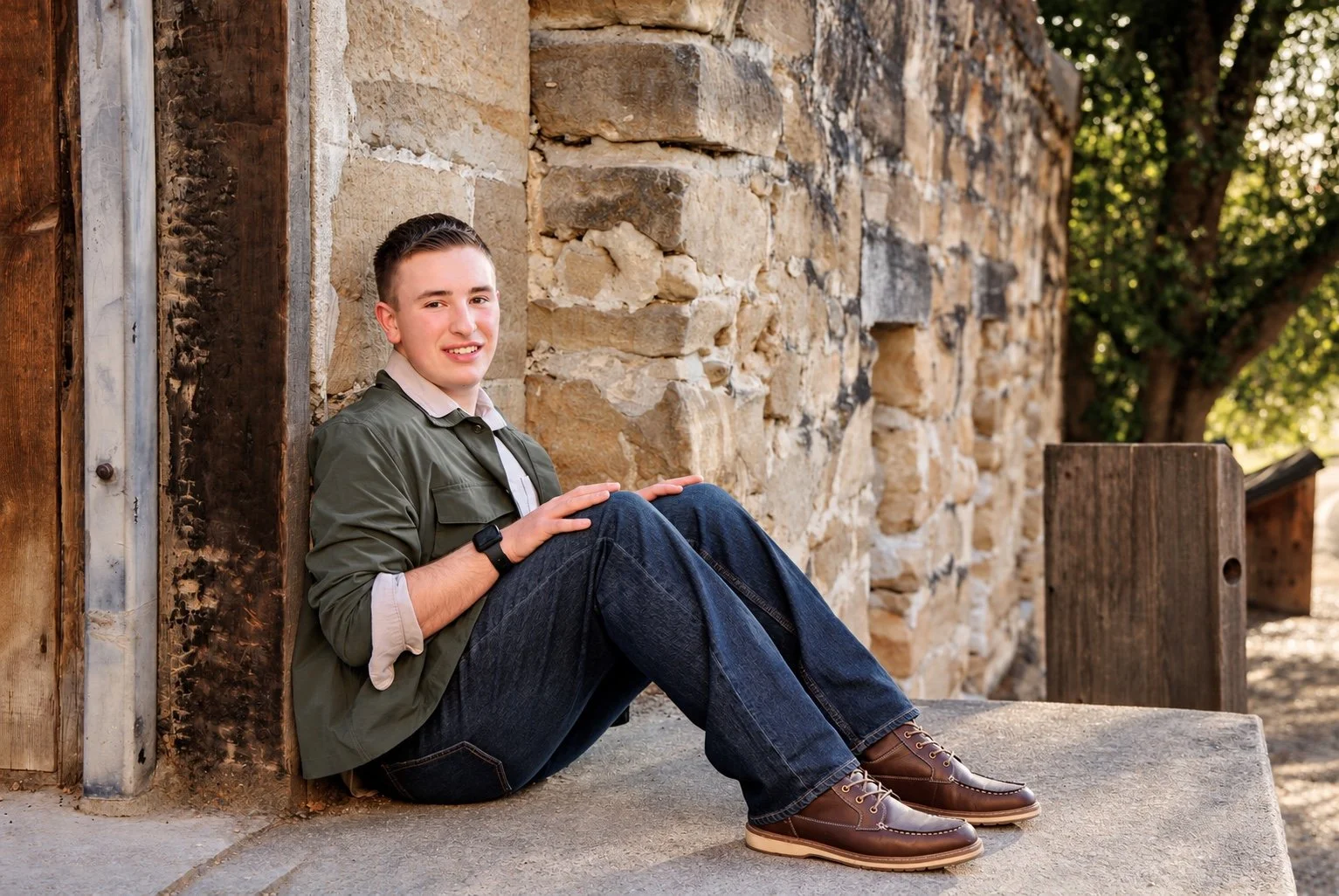 Man in a green shirt and jeans seated beside a rustic wooden door and stone wall, smiling at the camera.
