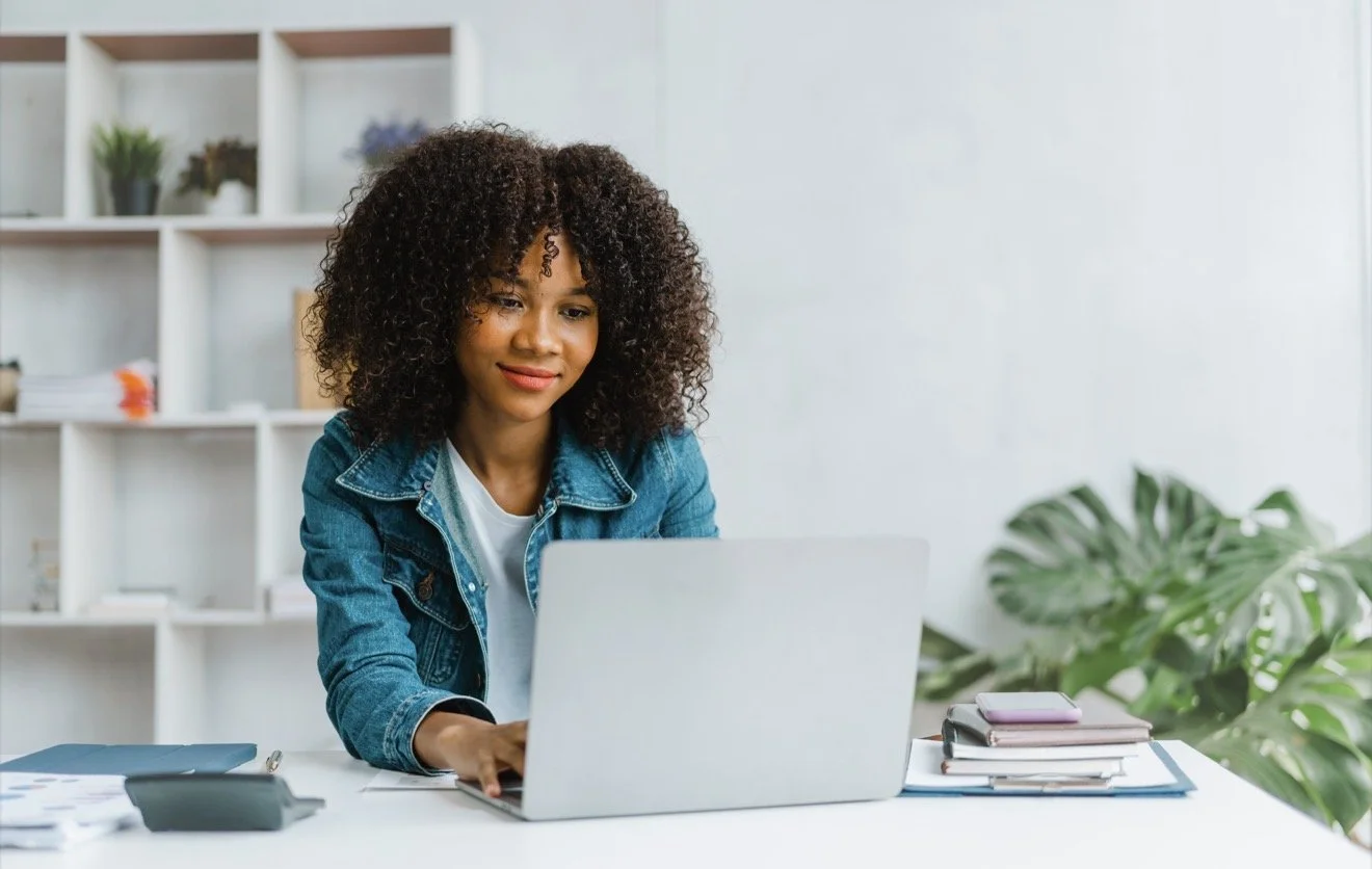 Woman in a denim jacket using a laptop at a desk beside books and a leafy plant.