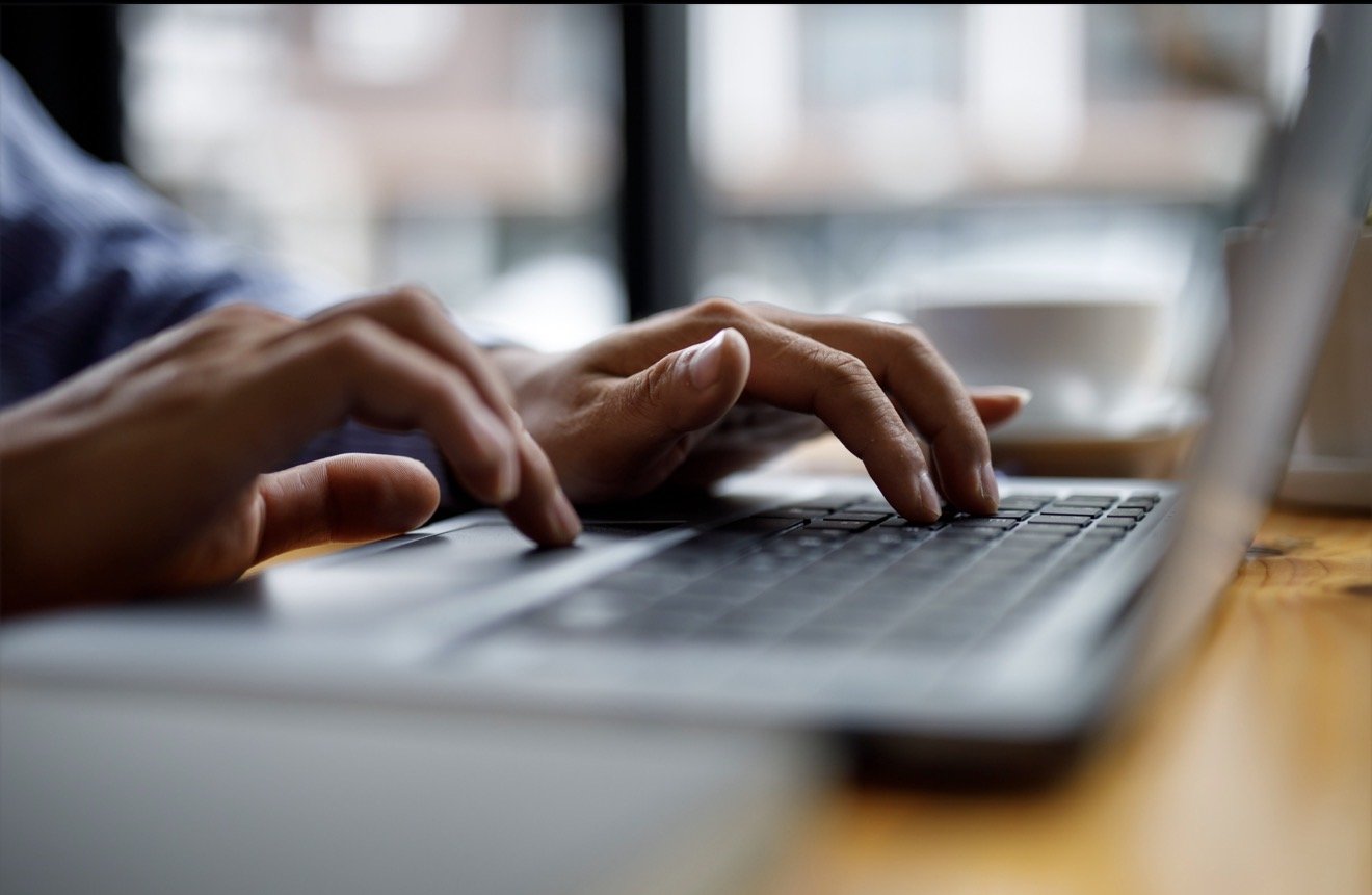 Close-up of hands typing on a laptop keyboard beside a window.