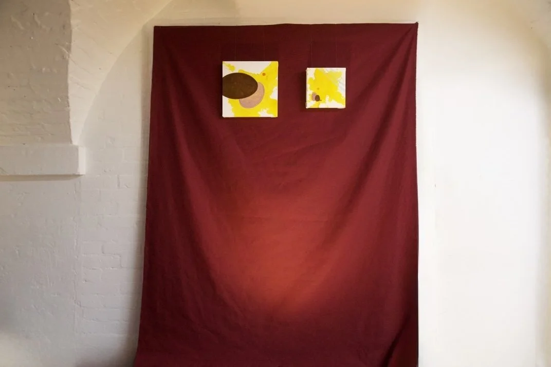 Two abstract paintings with yellow, brown, and beige shapes on white backgrounds, hanging on a red fabric backdrop.