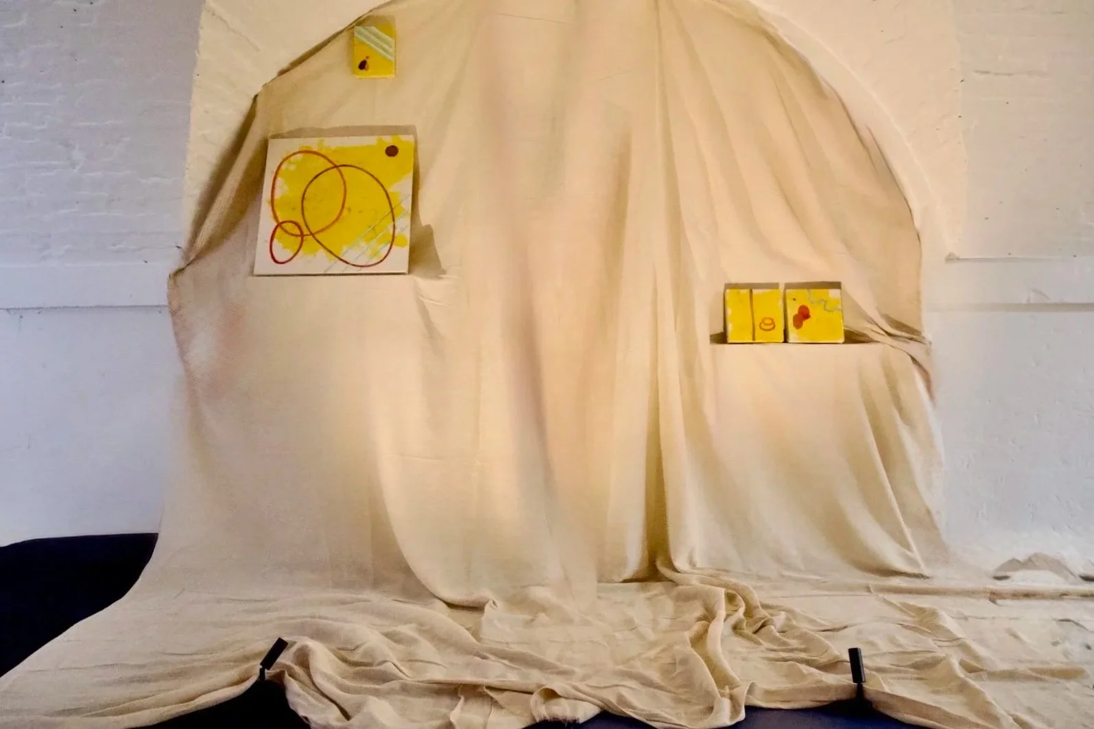 An art display with a cream-colored fabric backdrop, featuring two drawings on cardboard and three small painted canvases with abstract designs in yellow, red, and orange.