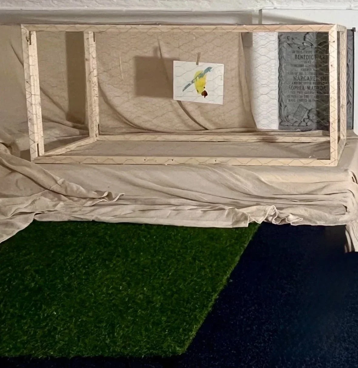 A wooden display case lying on a draped fabric surface, with a small illustrated card hanging inside. There is a black and green surface underneath the fabric.