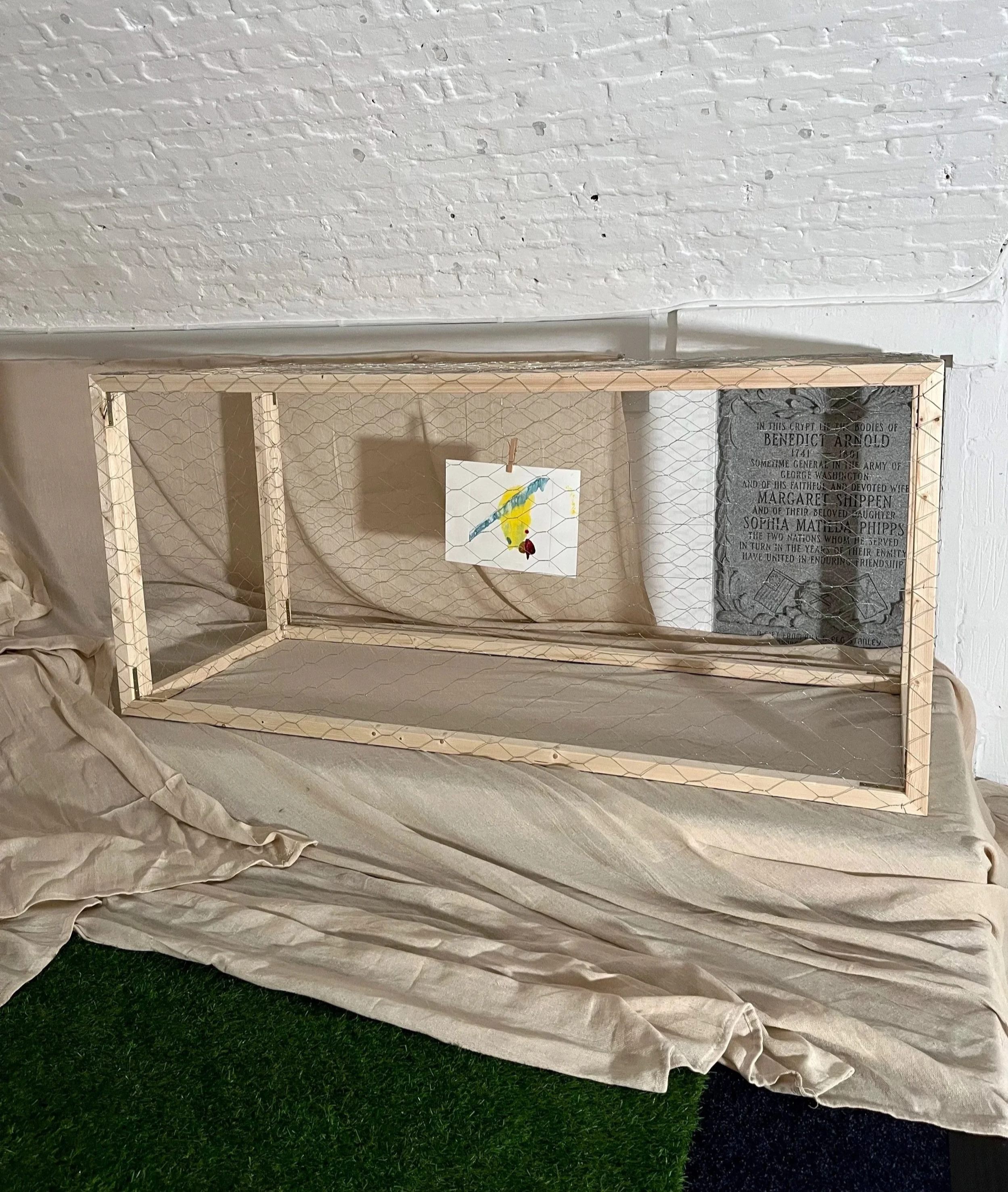 A rectangular wooden frame with chicken wire, placed on a beige cloth-covered surface against a white brick wall. Inside the frame, there is a small piece of paper with a watercolor painting of a bird and a red dot, hanging from a clothespin. To the right of the paper, a gray stone plaque with engraved text is mounted on the wall.