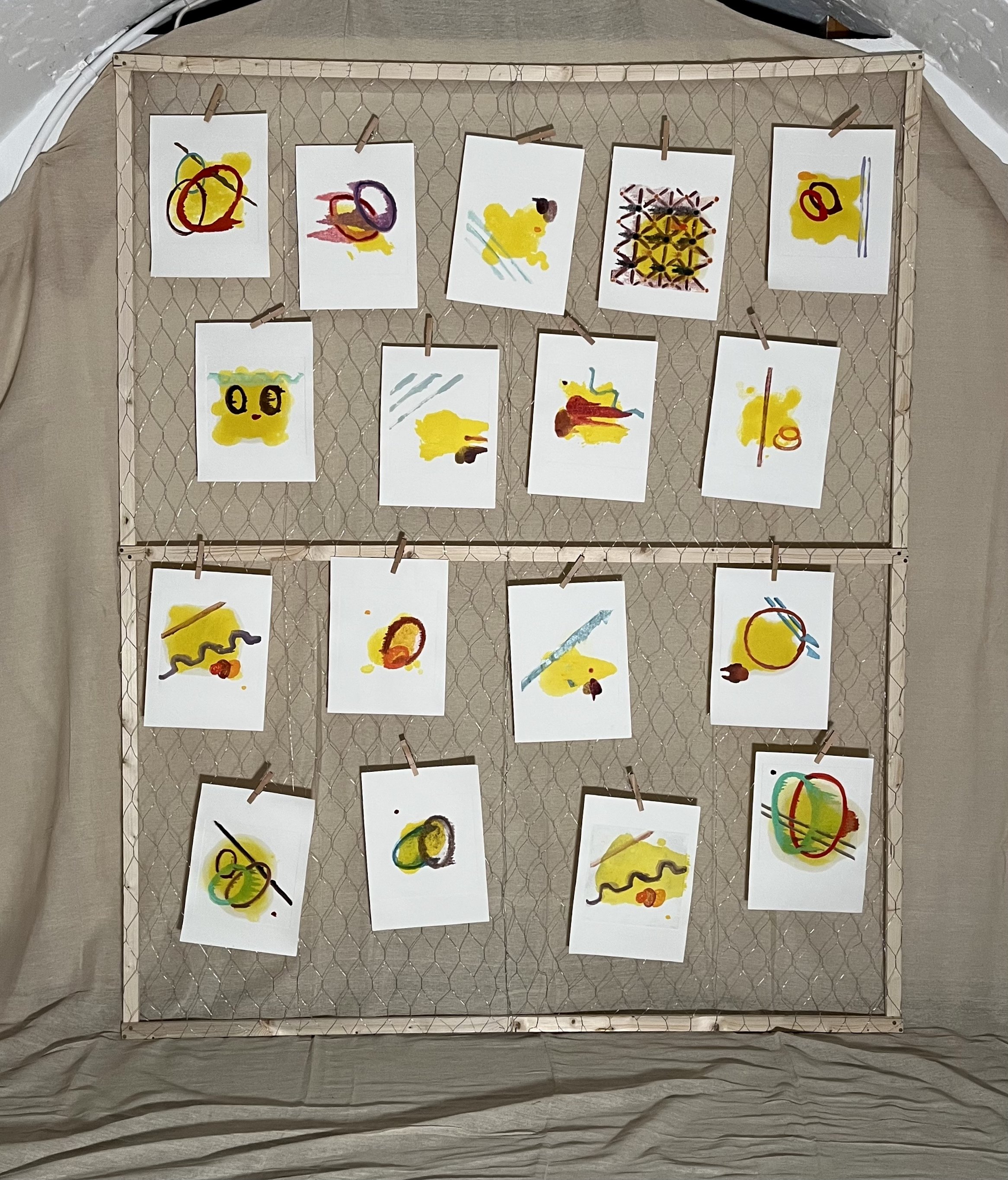 Collection of small abstract paintings with yellow, red, black, and blue brushstrokes hanging on a wire grid. Each artwork is fixed with small clothespins and arranged in a grid pattern.