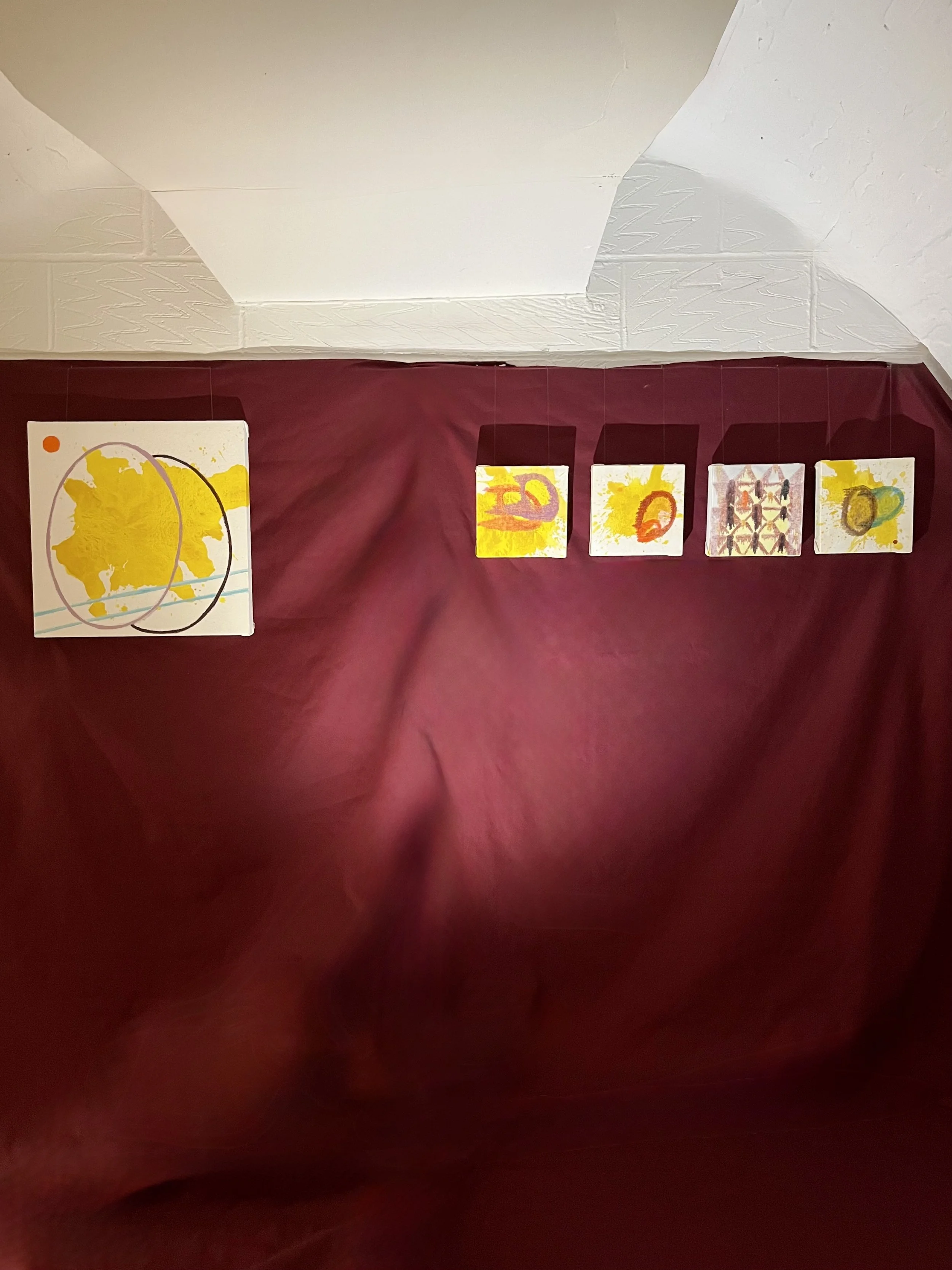 Abstract artwork featuring yellow, orange, and red shapes on white canvases, arranged on a dark red fabric background.
