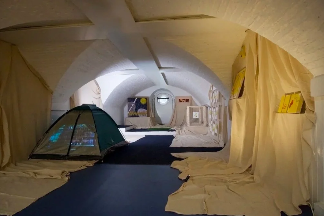 Indoor art installation resembling a cave interior with fabric drapes, a small tent, and abstract paintings.