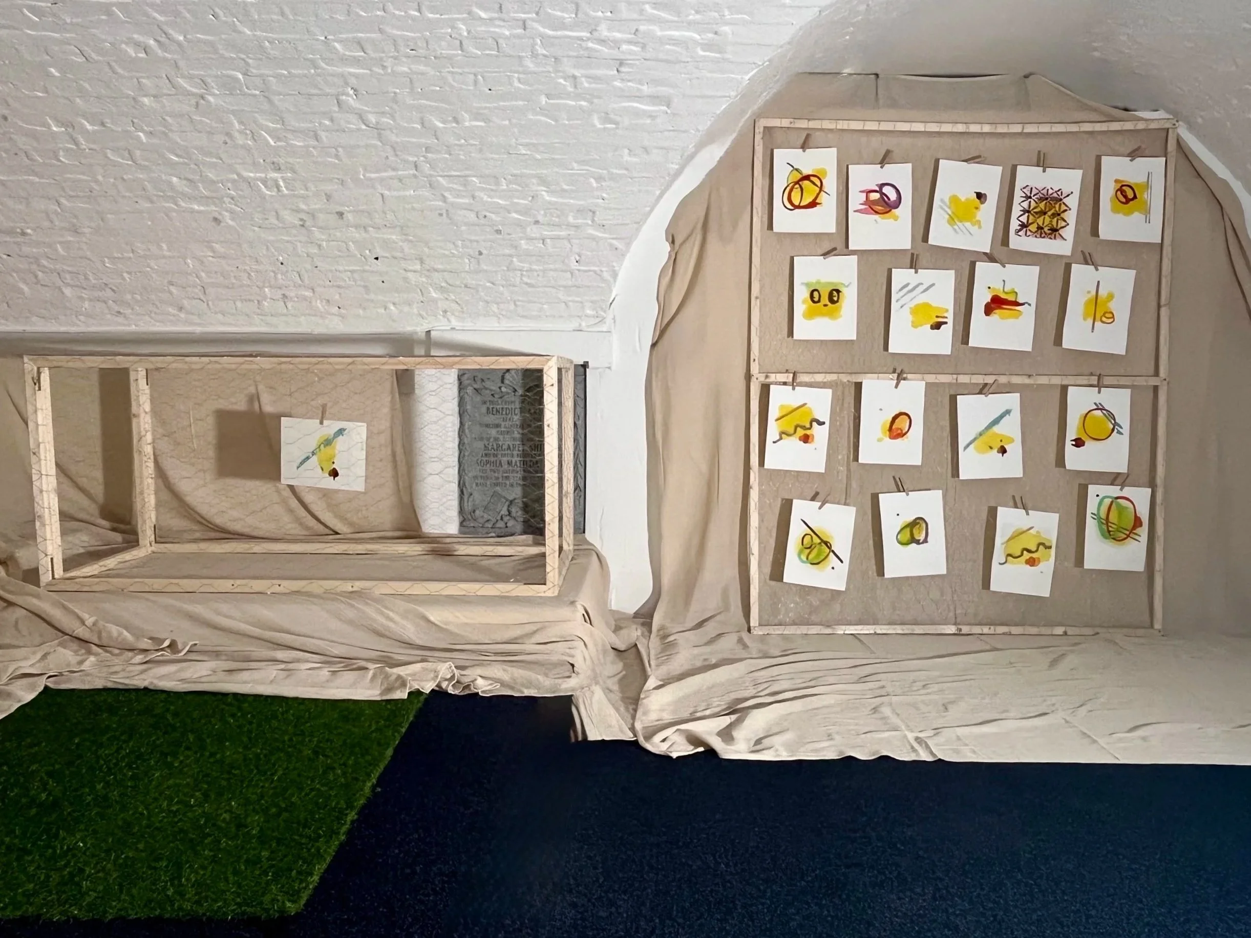 Display of abstract artwork on two fabric-covered panels with sketches attached by clothespins, set against a white brick wall and dark flooring.