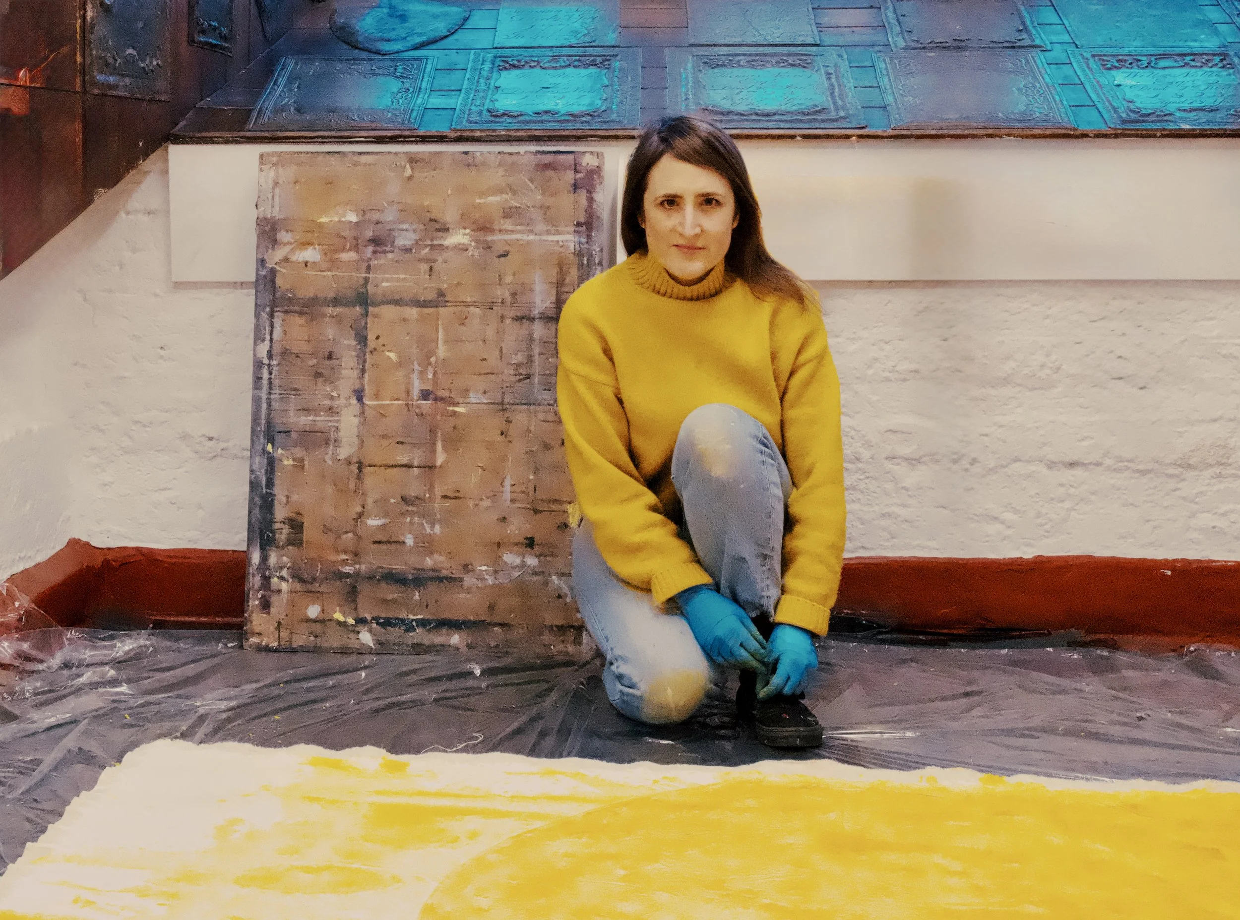 A woman wearing a yellow sweater and blue gloves, sitting on the floor and working on a painting in an art studio. There are paintings and art supplies around her.
