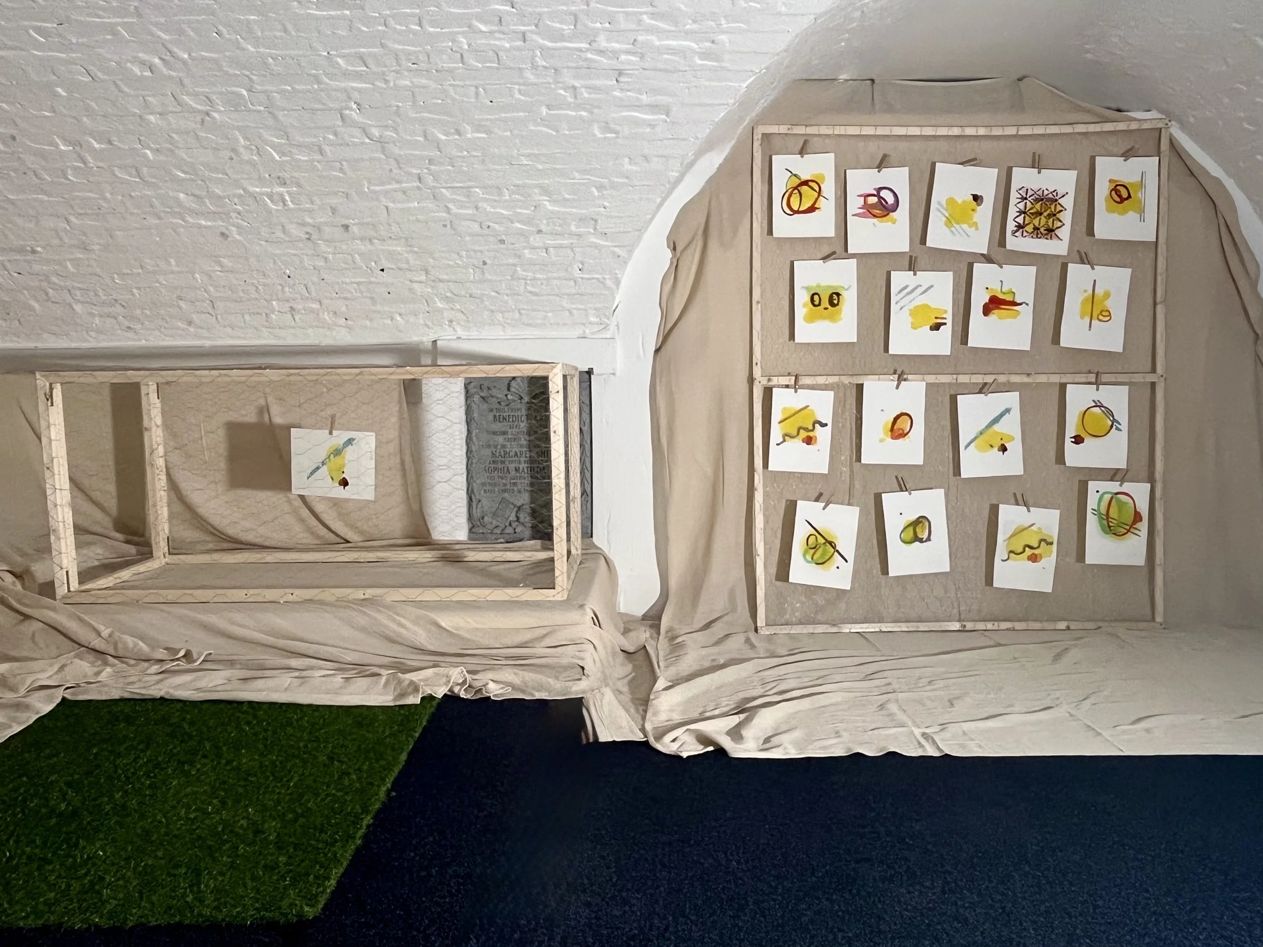 REFUGE at the Crypt exhibition display of abstract paintings arranged on a wire and wooden frame, draped cloth, set against a white brick wall and a chicken coop with artwork inside.